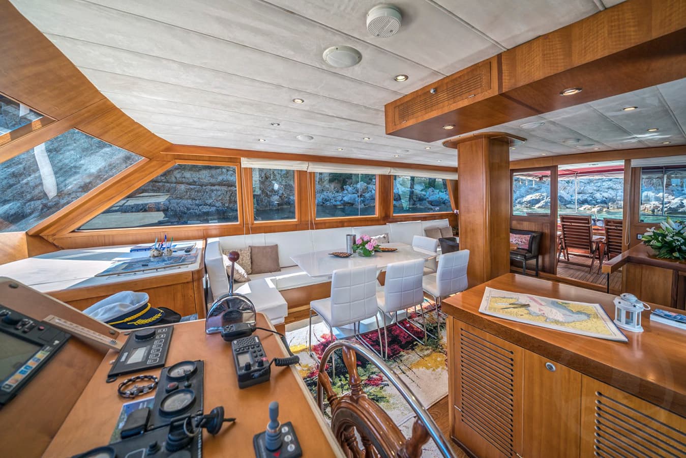 Yacht interior with helm station, white dining area, and rocky coastline visible through windows.