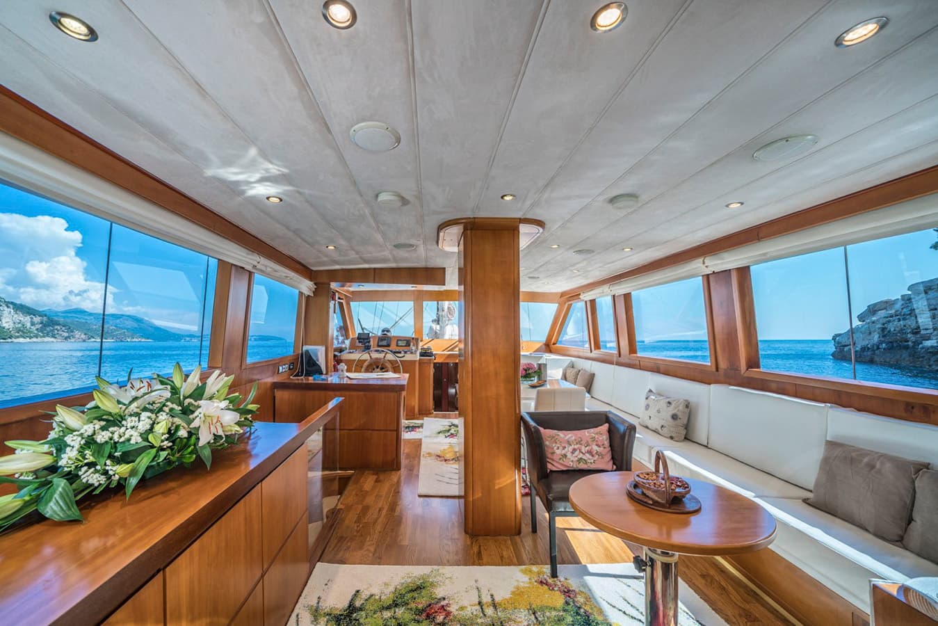 Yacht interior with wooden trim, white seating, and views of blue sea and rocky coastline.