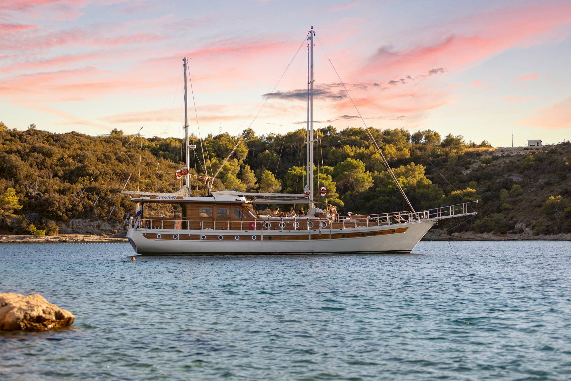 Wooden motor sailor anchored in calm blue water near a forested coastline at sunset.