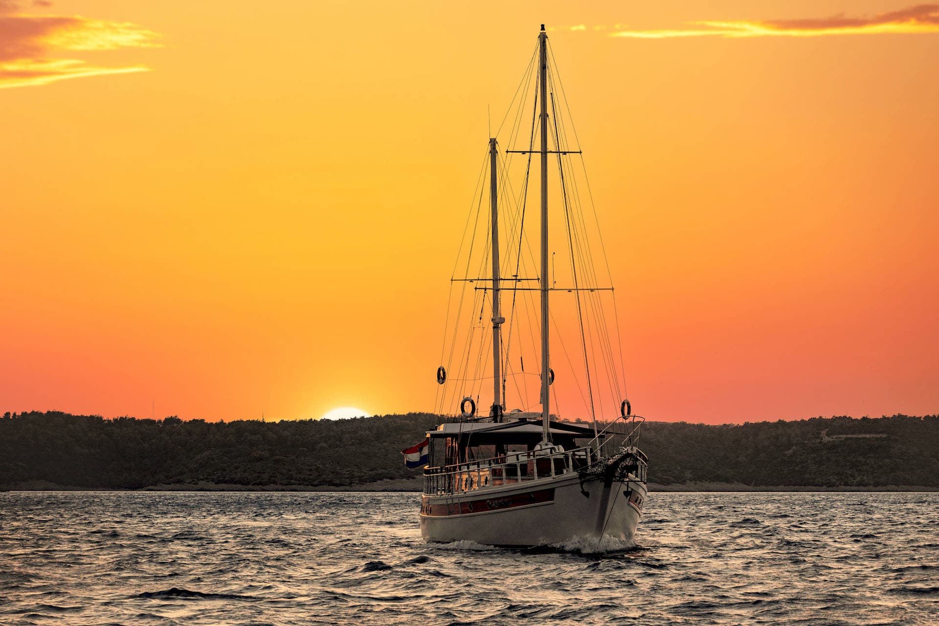 Sailboat cruising on choppy water against an intense orange sunset near a forested coastline.