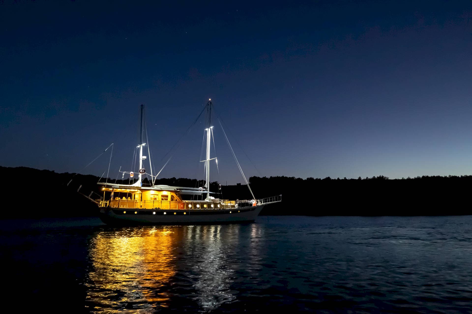 Illuminated wooden boat with masts on dark water at twilight near a forested shore.