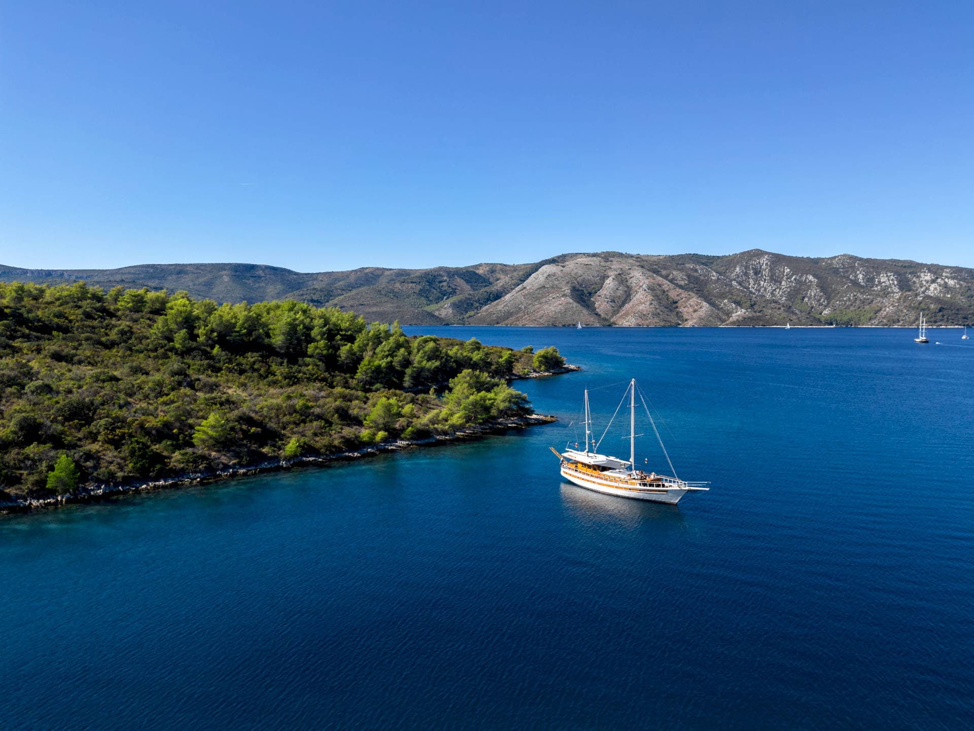Sailboat anchored in deep blue cove near forested, arid coastline under clear blue sky.