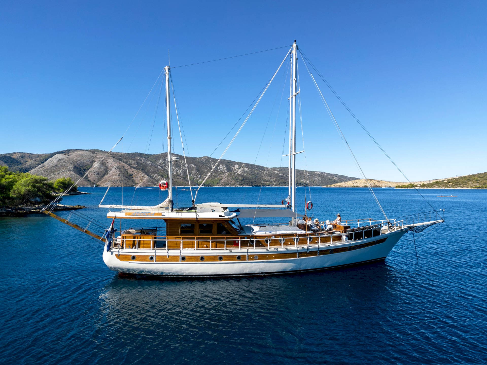 Wooden sailing yacht with passengers anchored in deep blue water near arid coastline