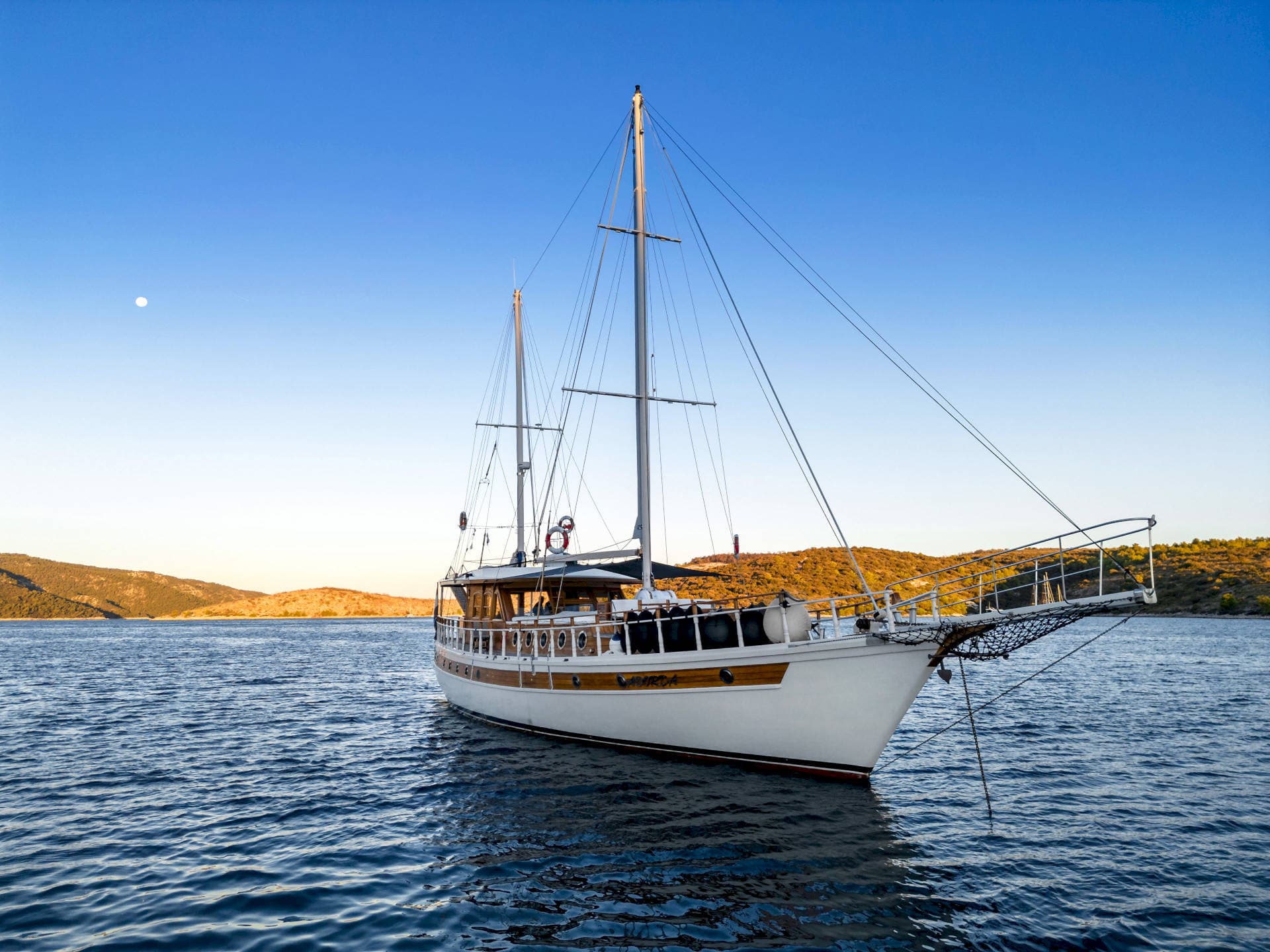 Large wooden sailboat anchored in deep blue water near arid, hilly coastline under clear sky.