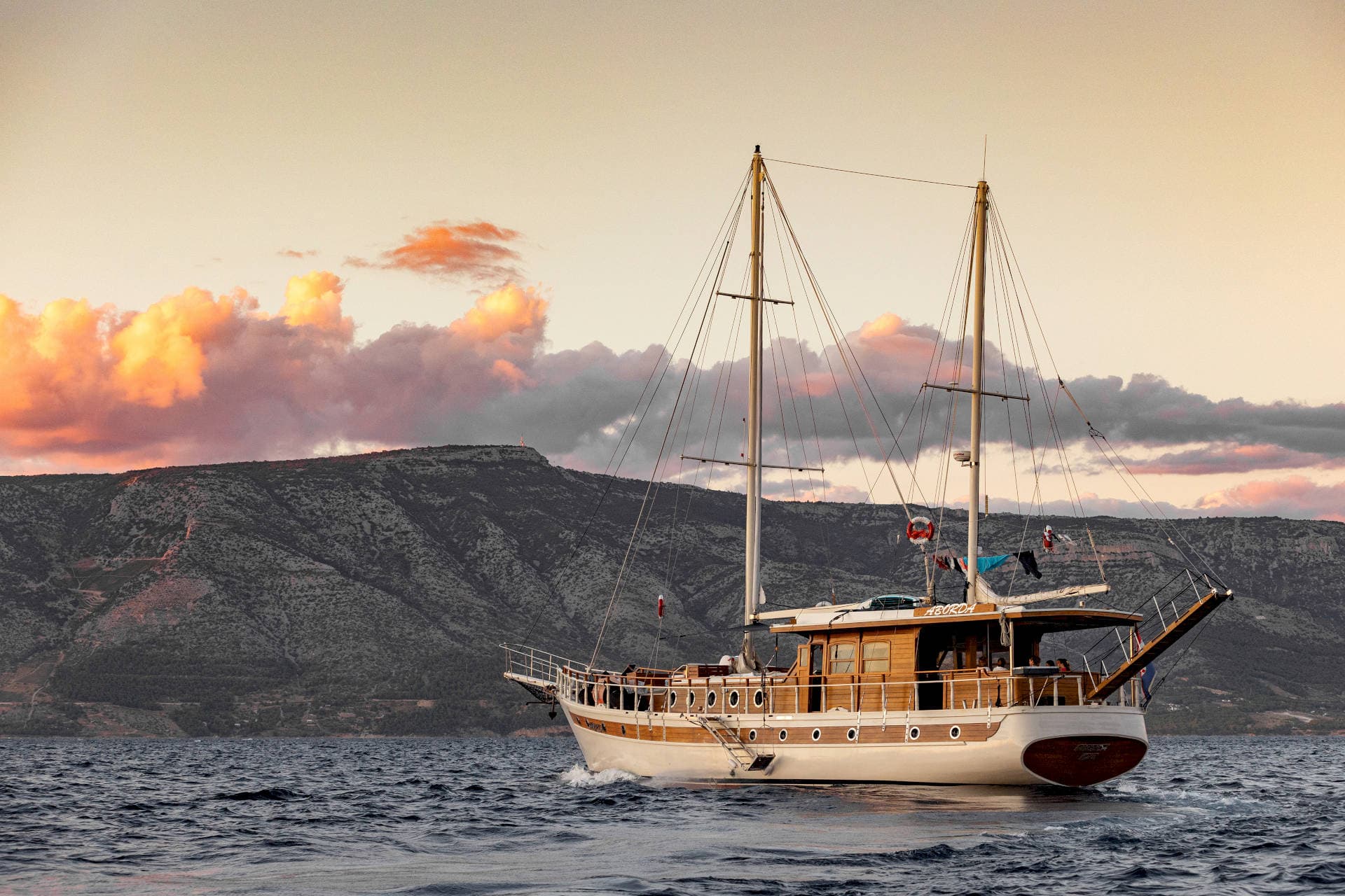 Wooden motor sailer boat cruising on dark water near a large, arid mountain range at sunset.