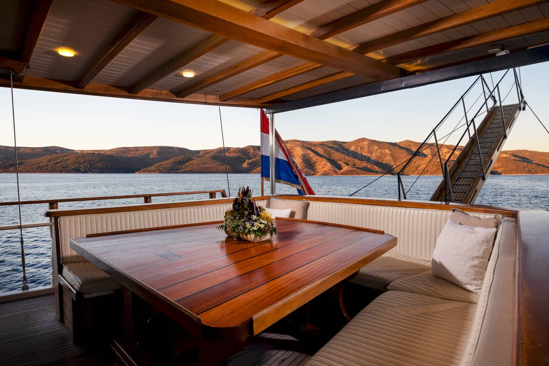 Boat dining area with wooden table, Croatian flag, and mountainous coastline view.