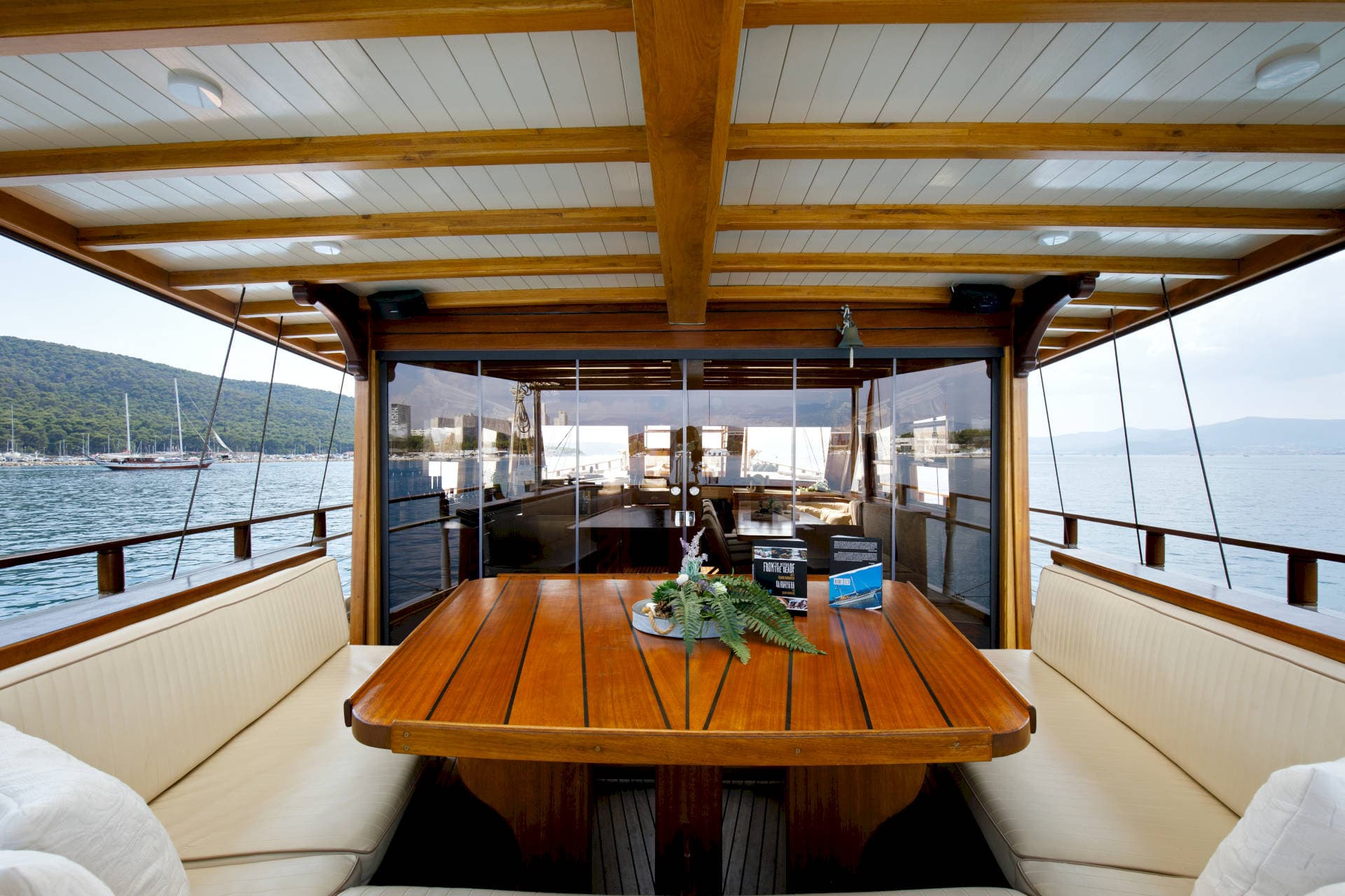 Wooden deck dining area on a boat overlooking a harbor with forested hills.