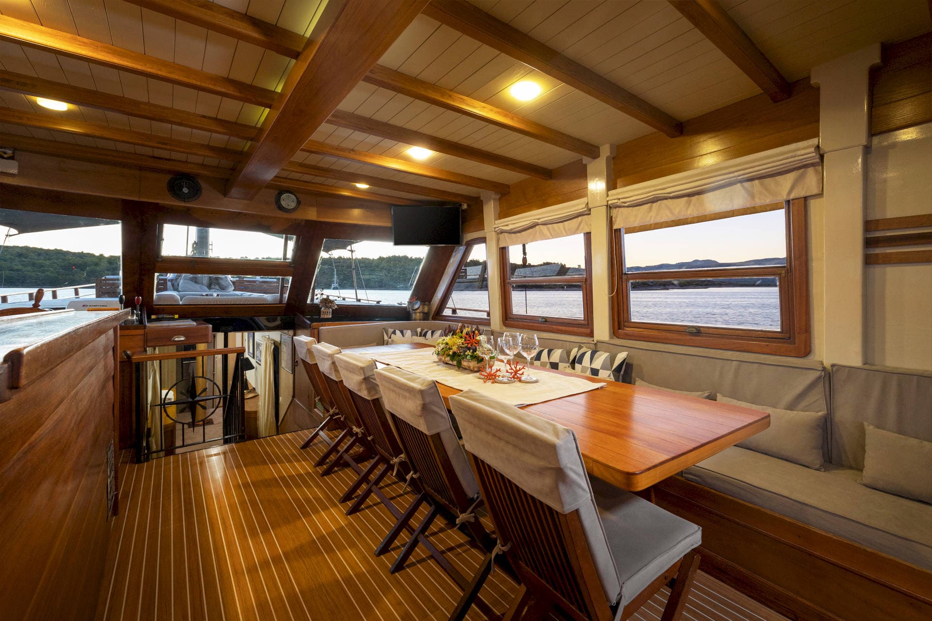 Wooden yacht dining area with set table overlooking calm water and distant hills at sunset