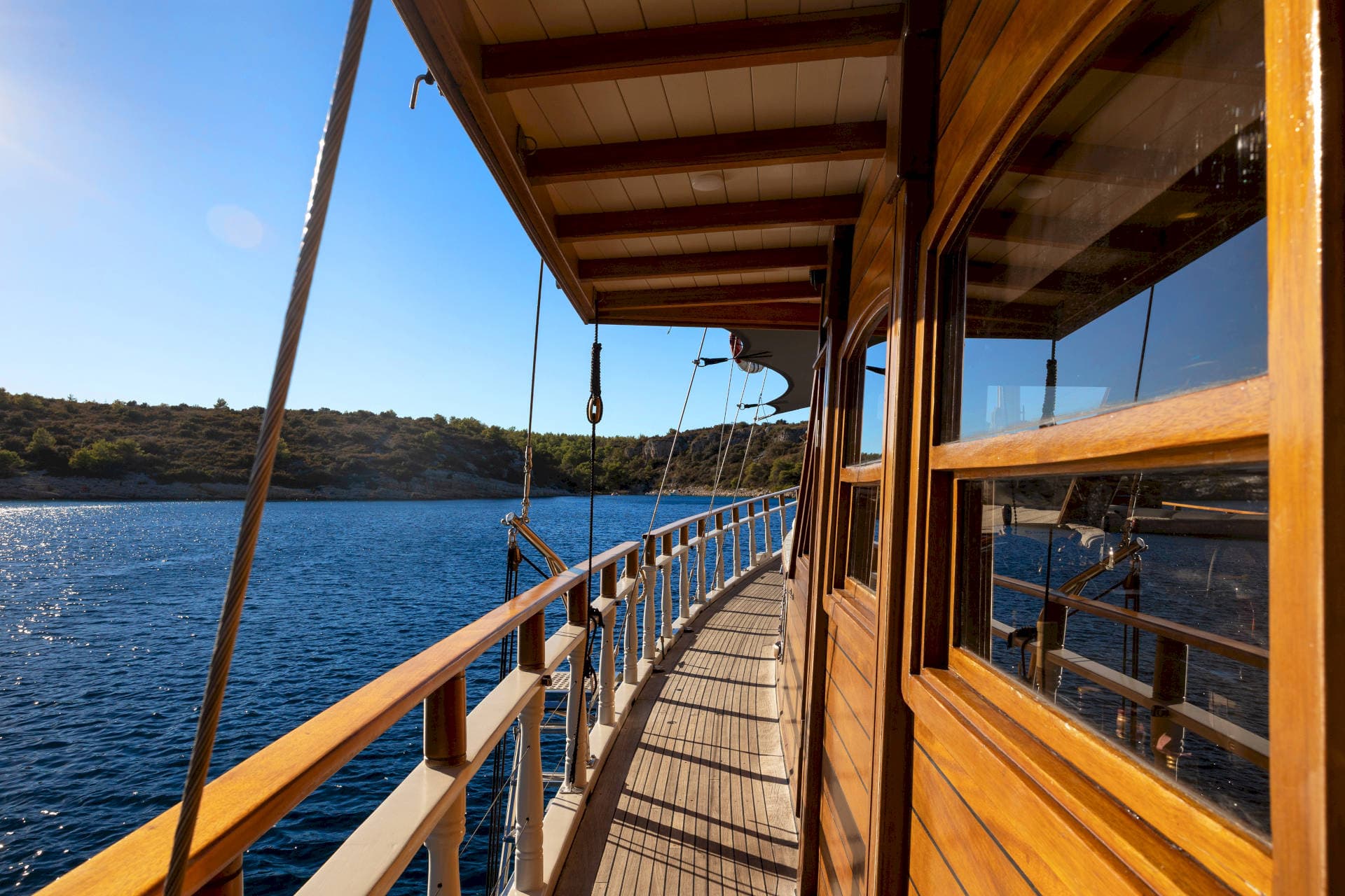 Wooden deck and cabin side of a boat sailing past a green, scrub-covered coastline.