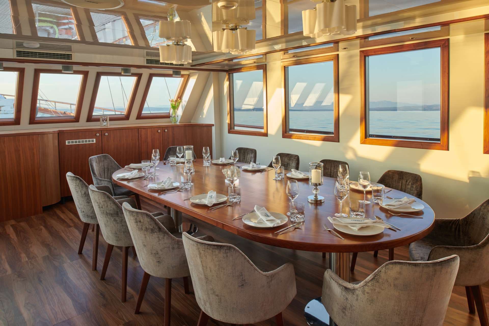 Dining room aboard a vessel with a long wooden table set for dinner overlooking the sea.