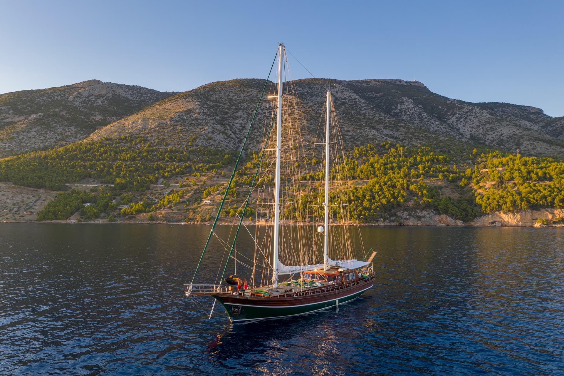 Wooden sailboat anchored on dark blue water near a steep, scrub-covered coastline.