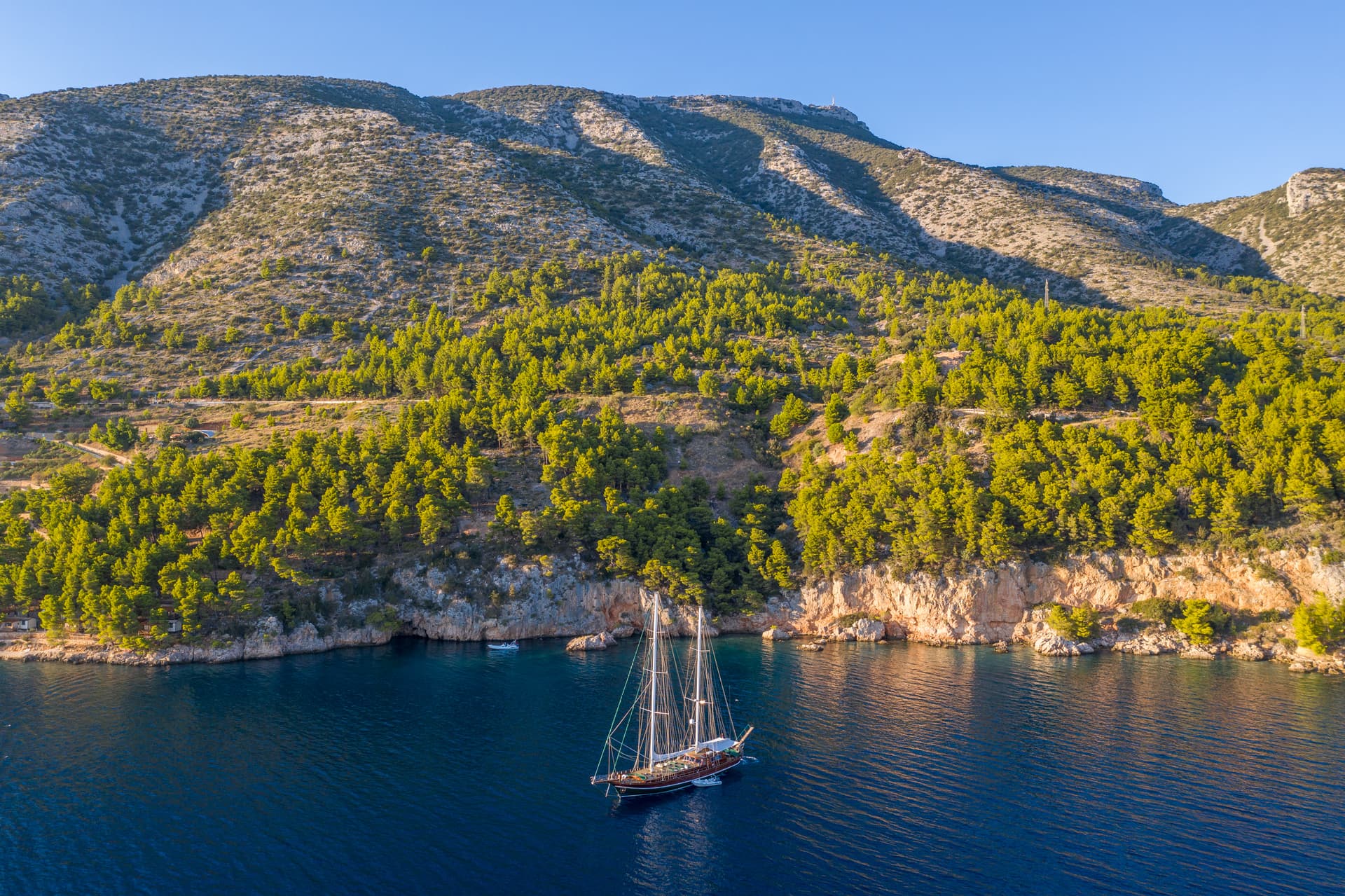 Large sailboat anchored in deep blue cove below steep, sunlit, scrub-covered mountains.