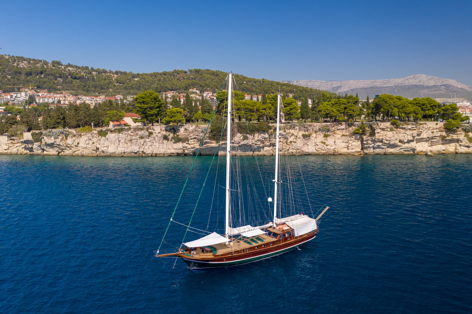 Wooden sailboat anchored in deep blue water near a rocky, tree-covered coastline under a clear sky.