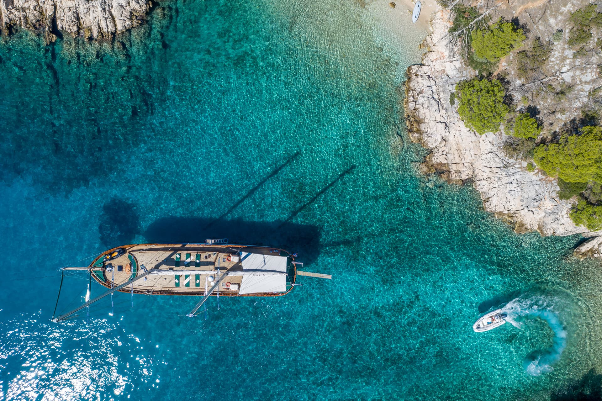 Aerial view of large wooden boat anchored near rocky coastline with turquoise water and small speedboat.
