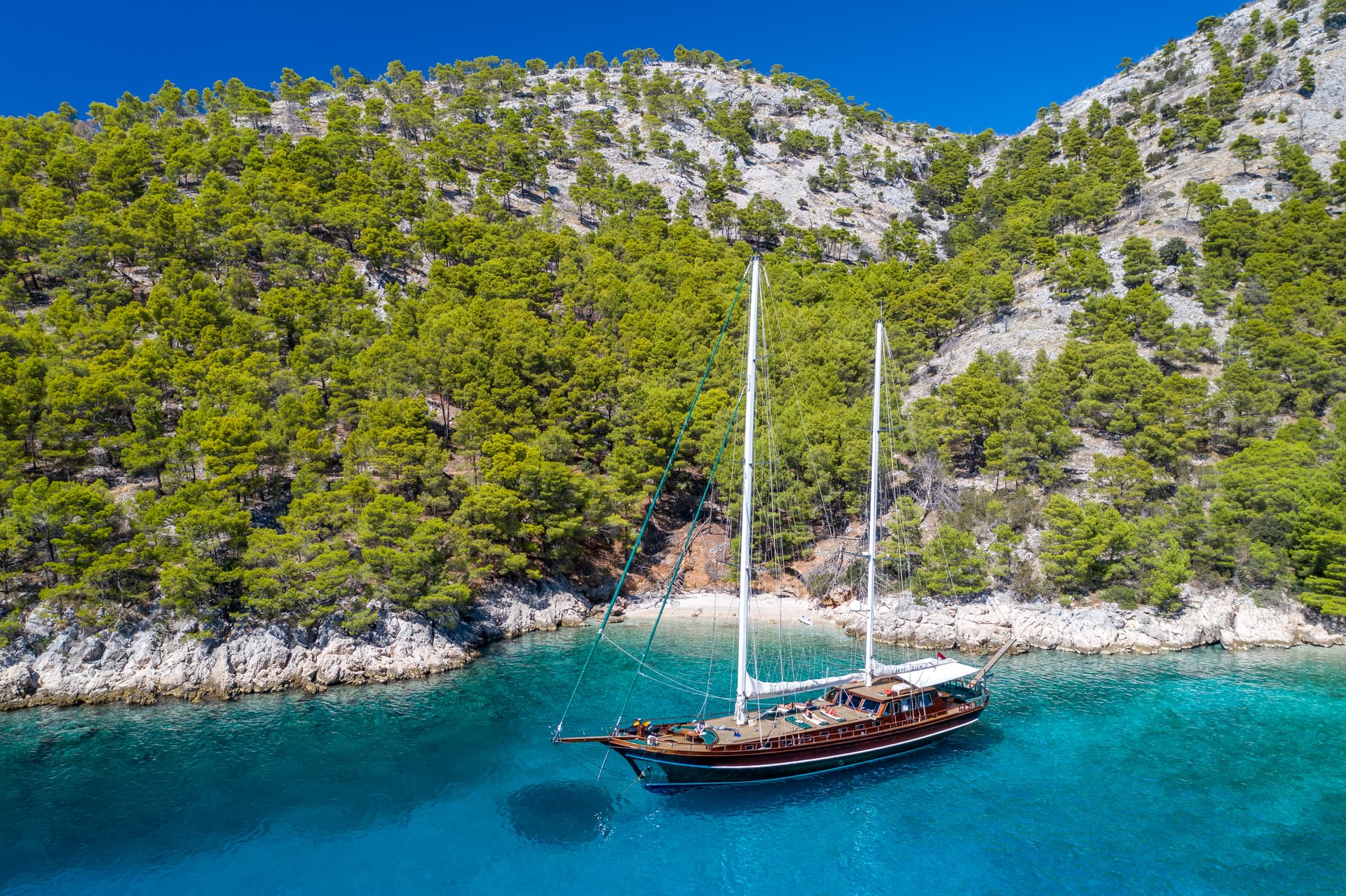 Wooden gulet sailboat anchored in turquoise cove below pine-covered rocky hillside
