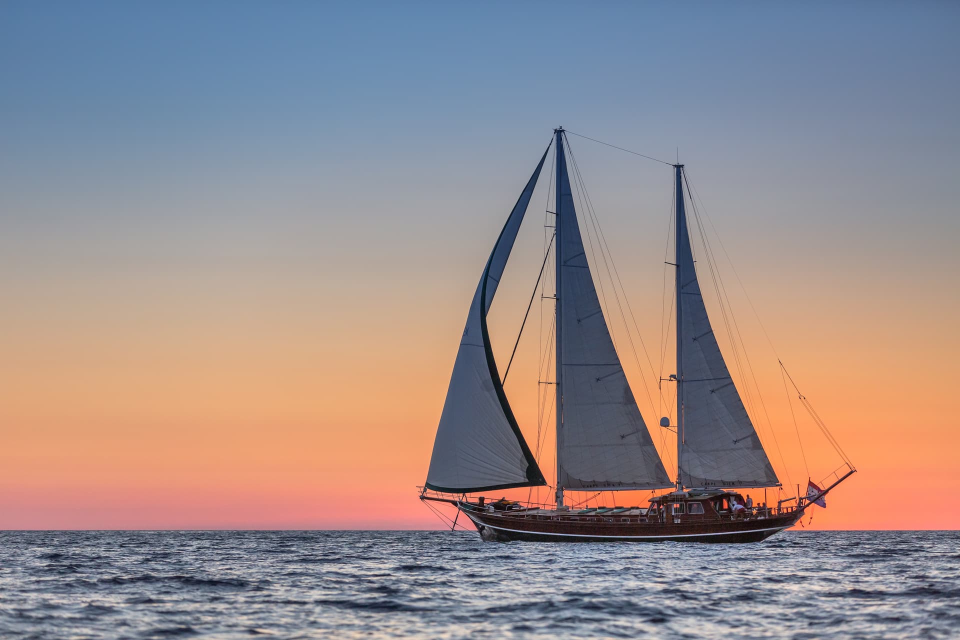 Two-masted wooden sailboat with sails up on choppy water against an orange and blue sunset.