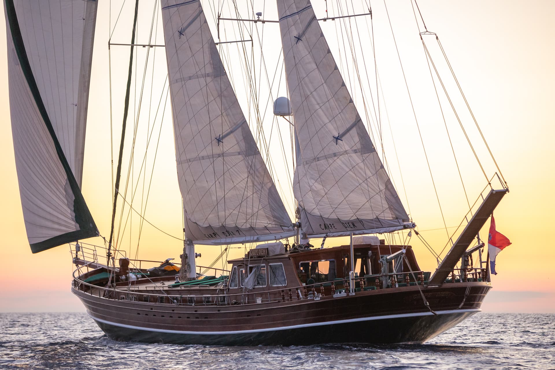 Wooden sailboat with sails up on wavy water against a bright sunset sky