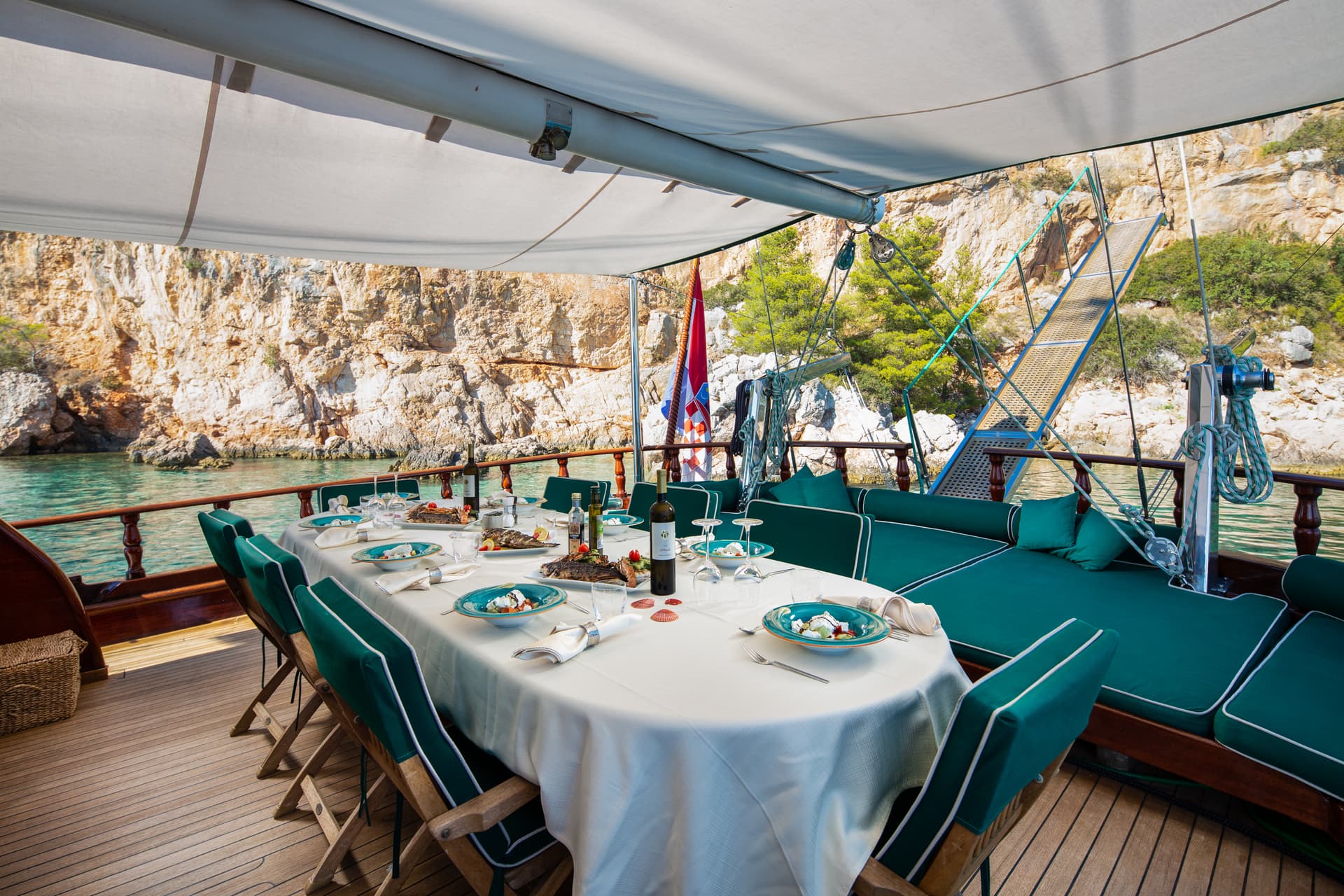Dining table set for a meal on a boat deck next to a rocky, green coastline.