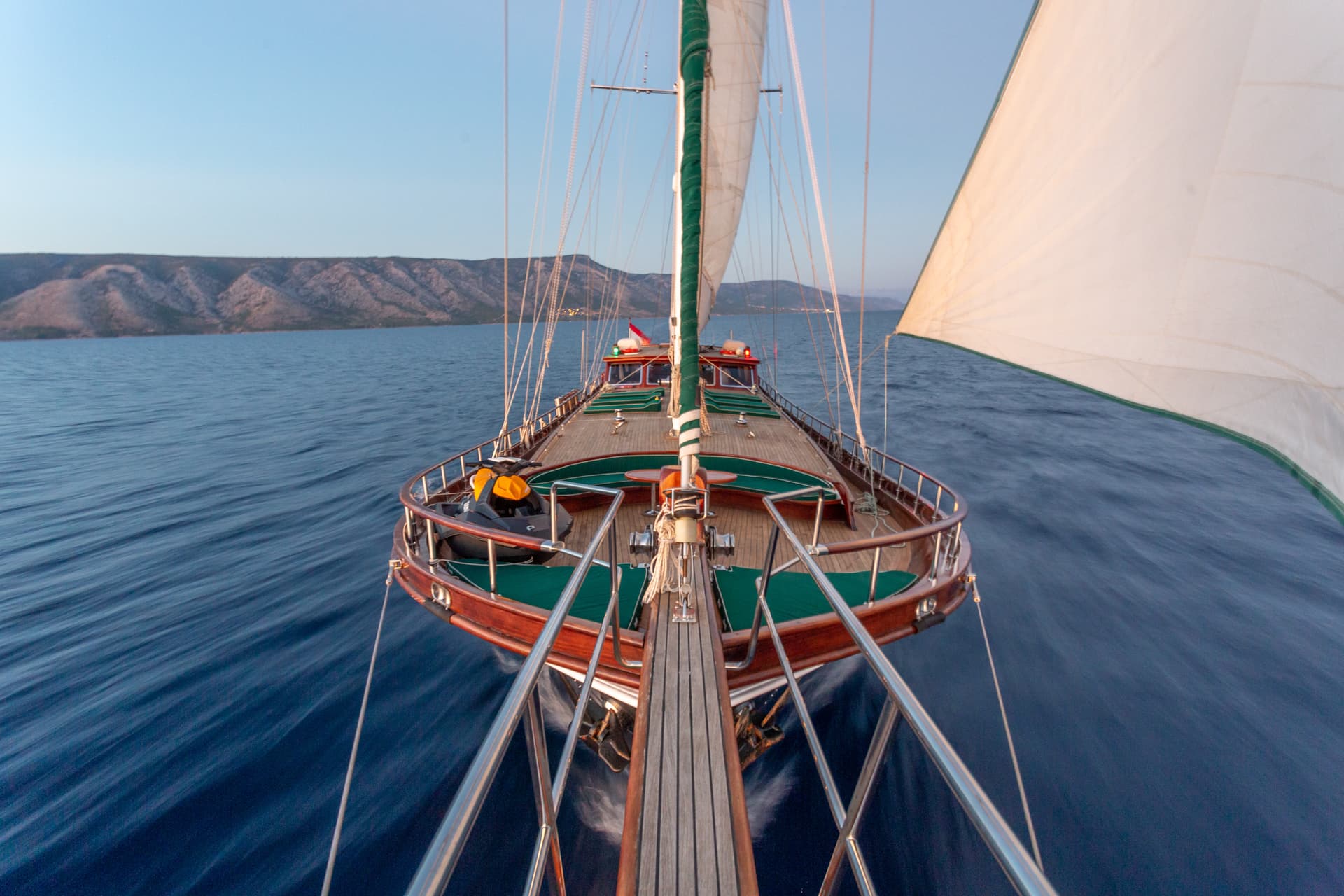 Wooden sailboat with sails up moving across dark blue water near mountains.