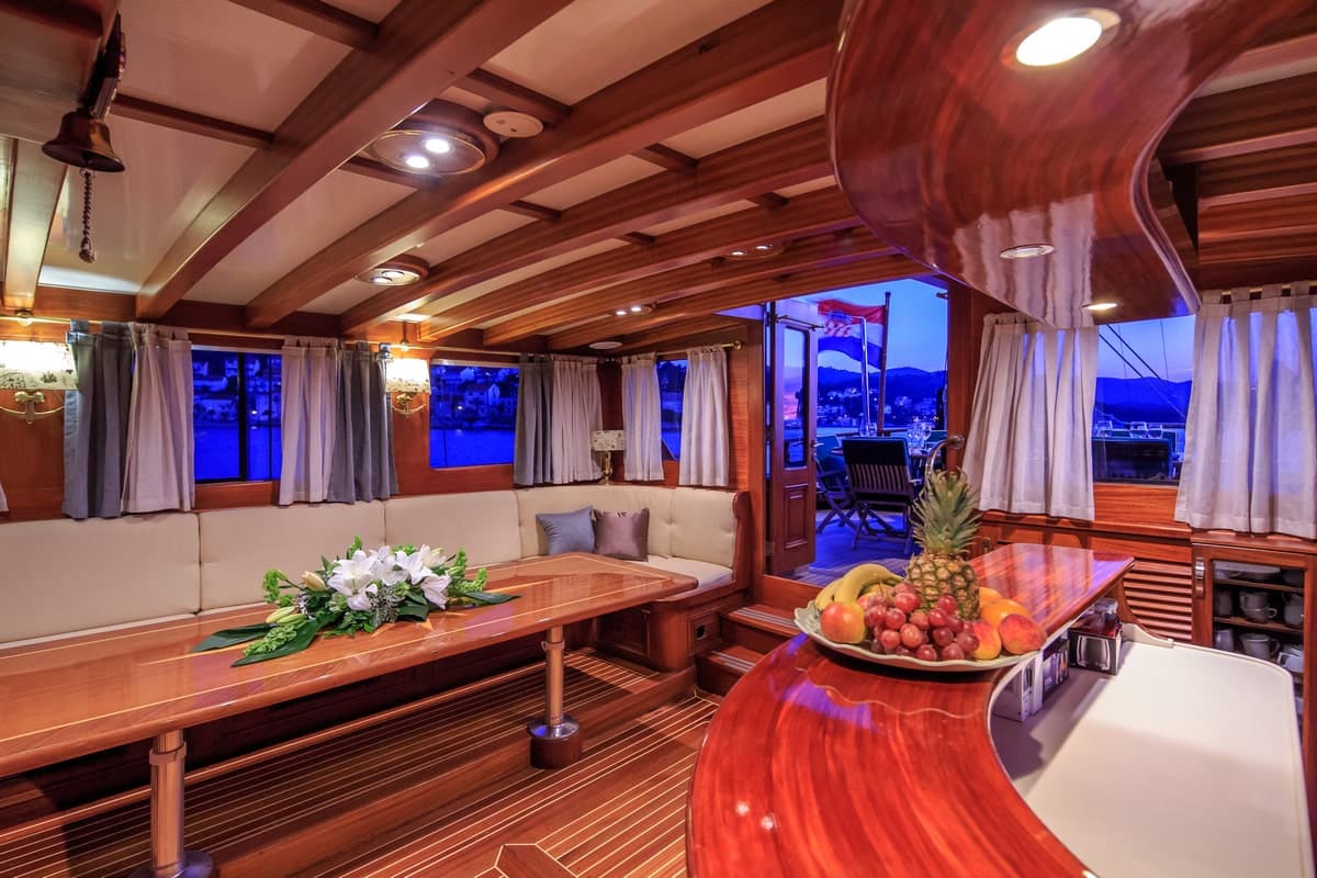 Yacht interior with polished wood, dining area, and fruit bowl overlooking a harbor at dusk.