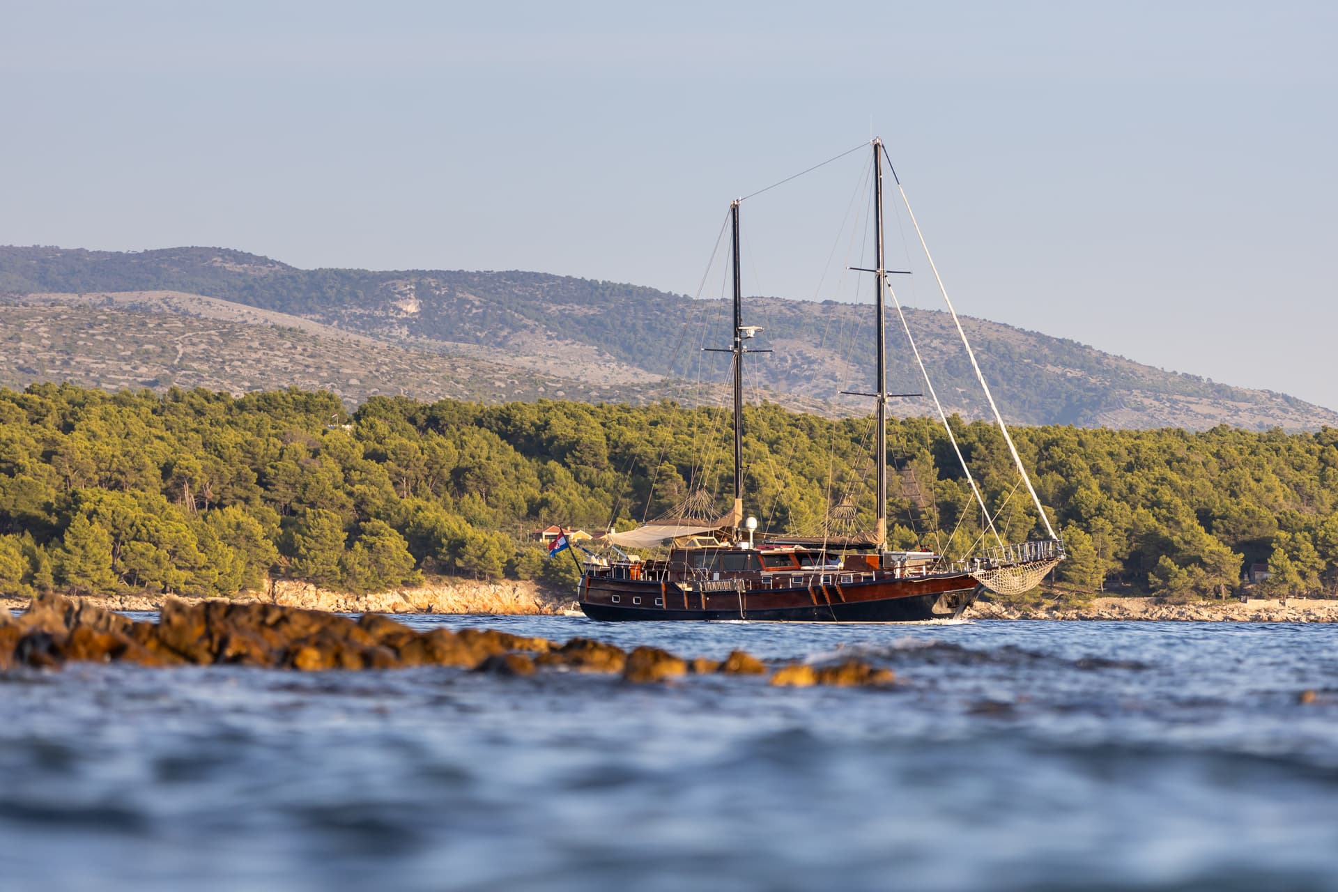 Wooden sailboat near forested coastline with dry hills under clear sky