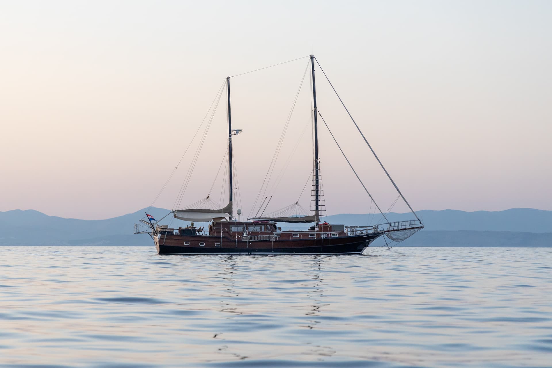 Wooden sailboat anchored on calm water with hazy mountains at sunset