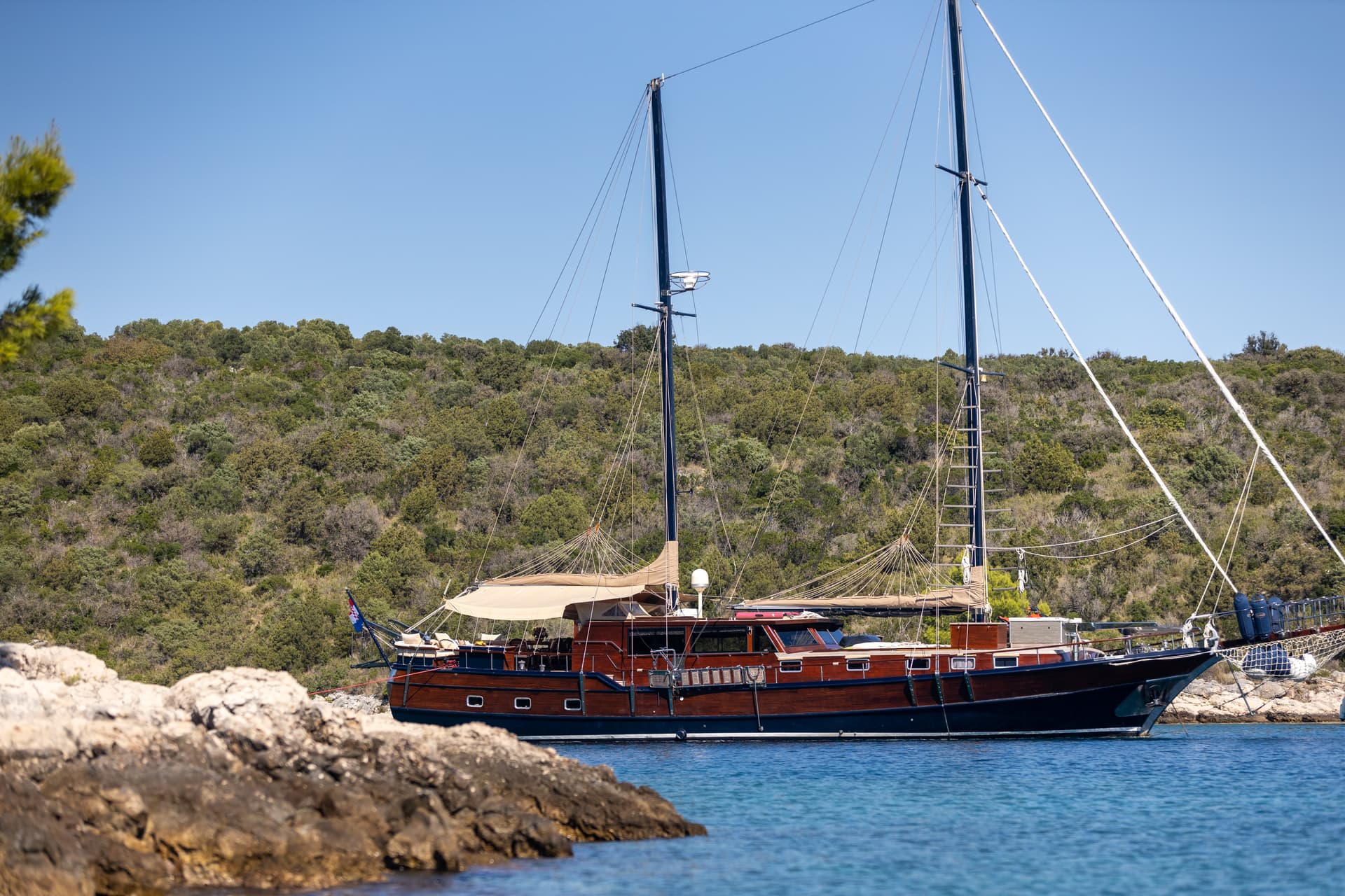 Wooden sailboat moored in clear blue water near a rocky, scrub-covered coastline.