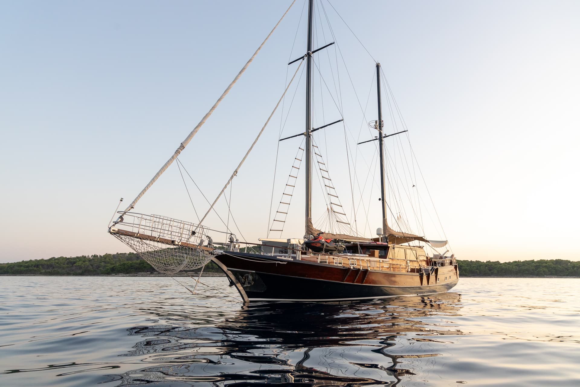 Large wooden sailboat anchored on calm water near a forested coastline at sunset.