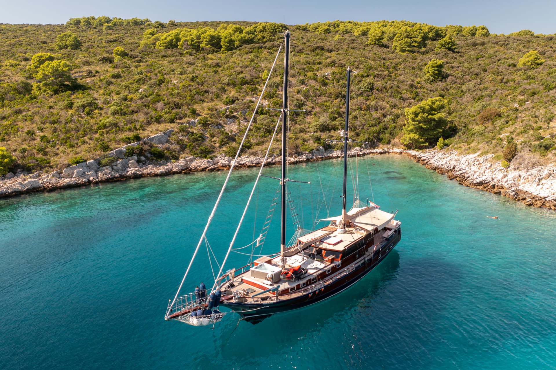 Large wooden sailboat anchored in turquoise cove next to scrub-covered rocky coastline.