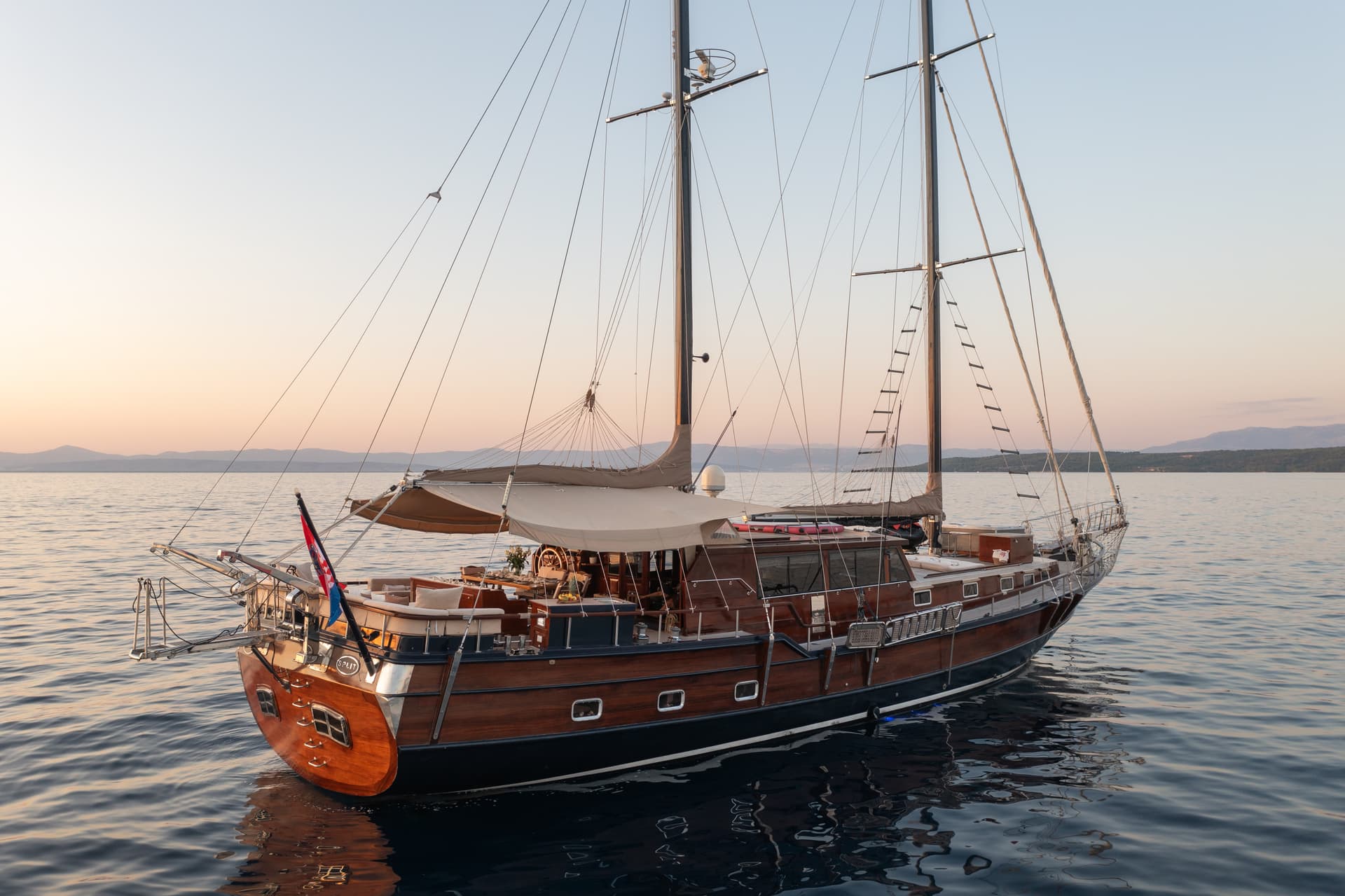 Wooden gulet yacht anchored on calm water near distant mountains at sunset, Croatian flag visible.
