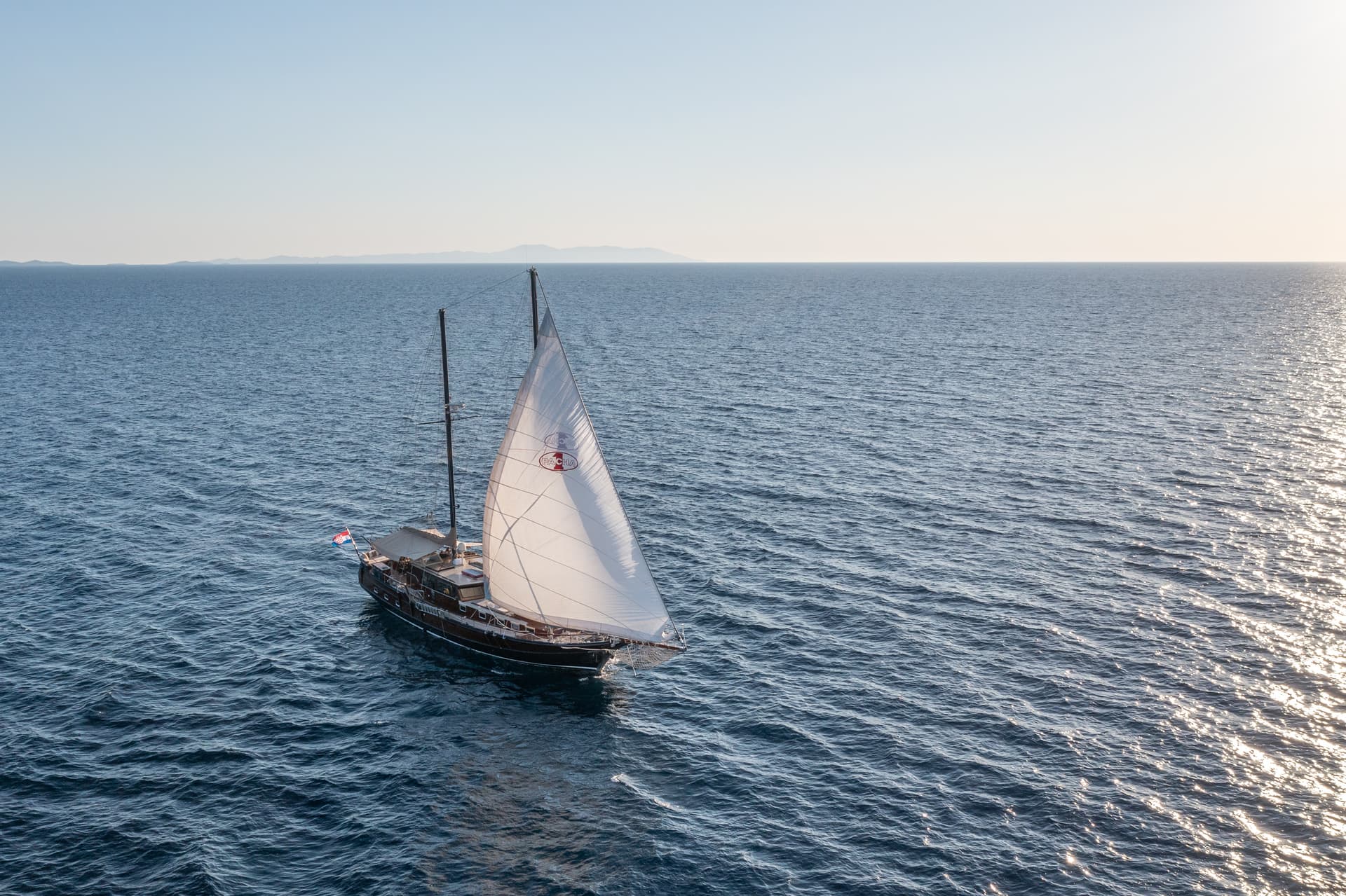 Sailboat with white sail on deep blue, sun-dappled sea with distant islands visible.