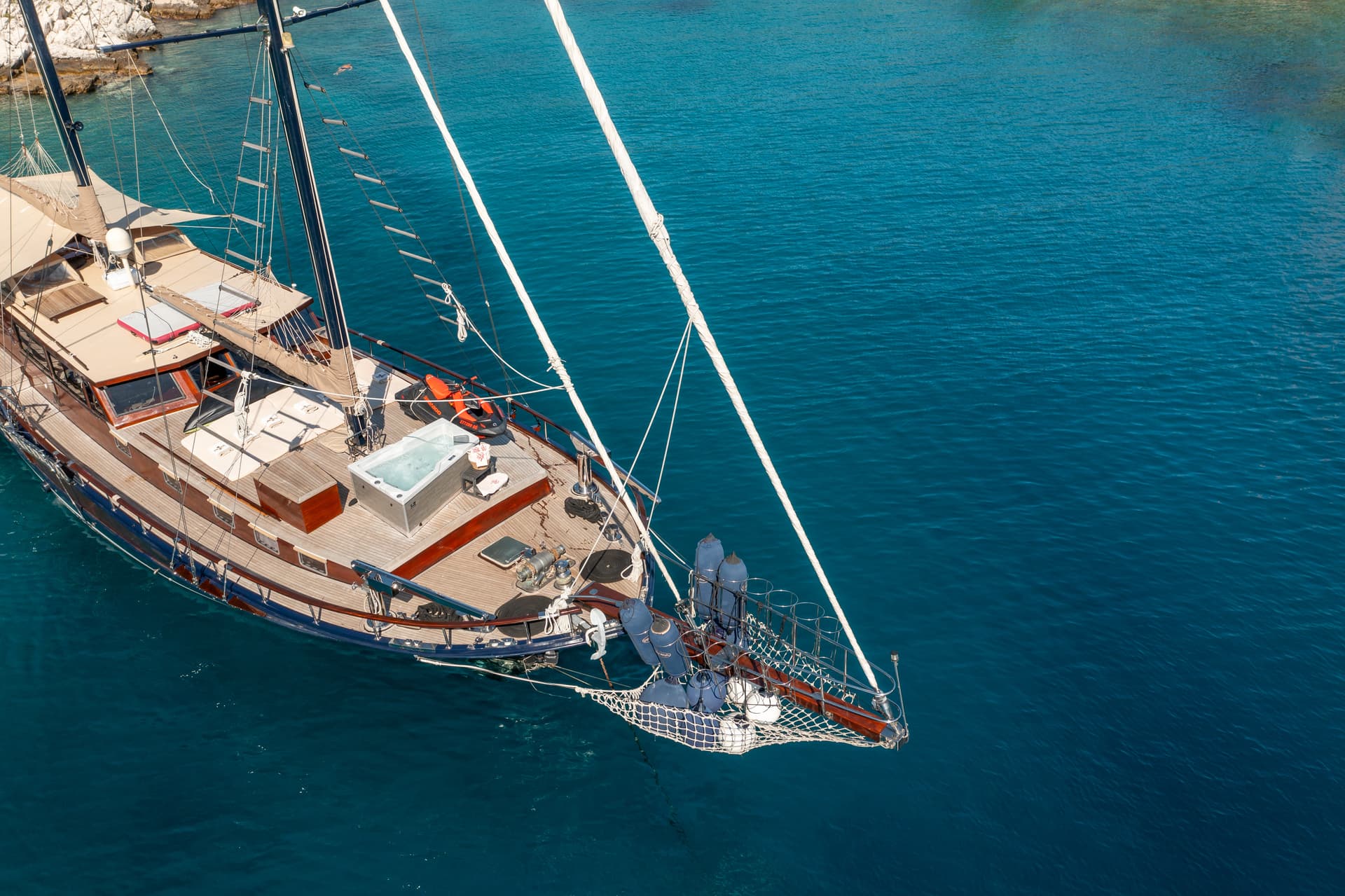 Luxury wooden sailboat with hot tub and jet ski anchored in clear blue coastal water.