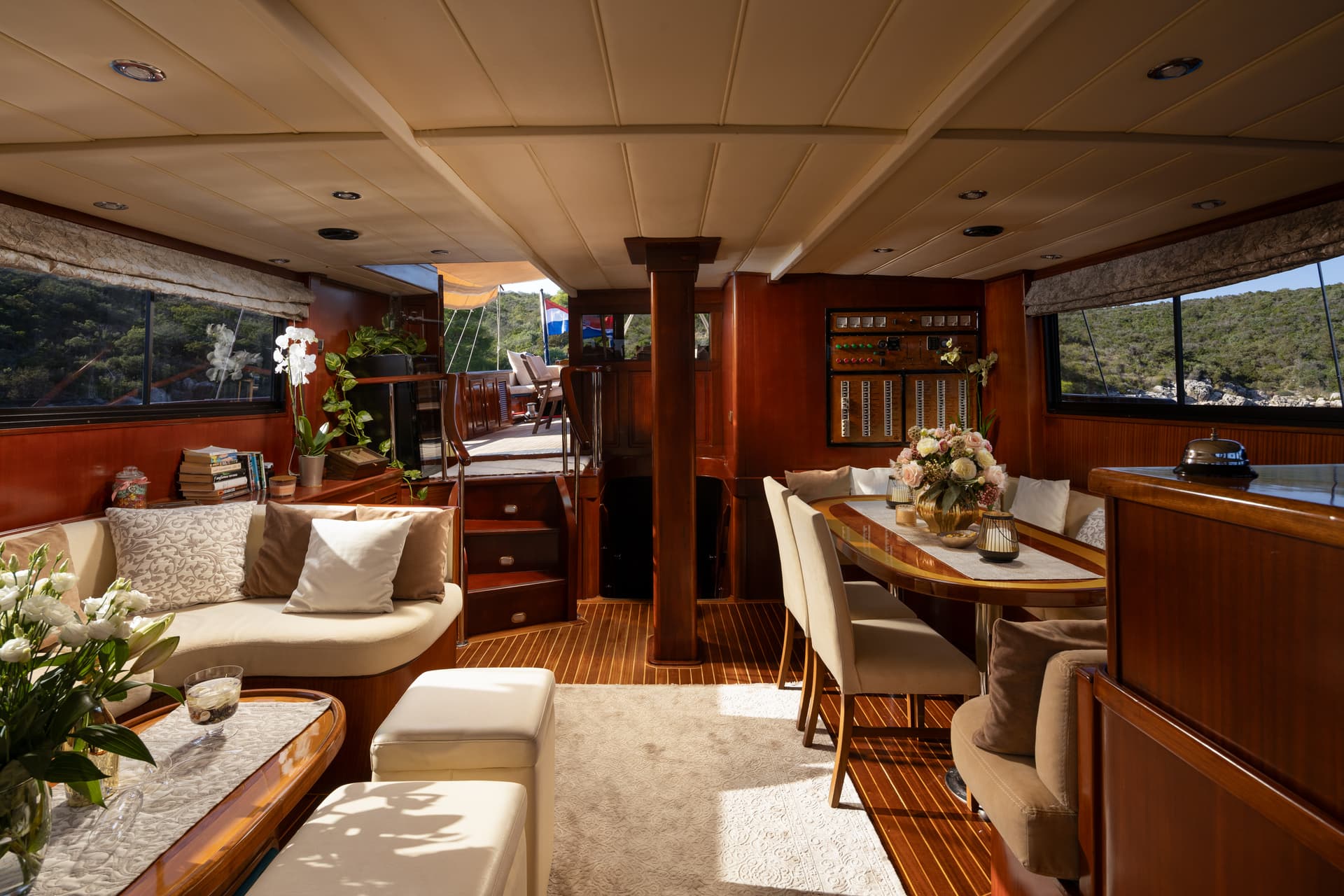 Interior salon of a yacht with wood paneling, dining area, and view of green coastline.