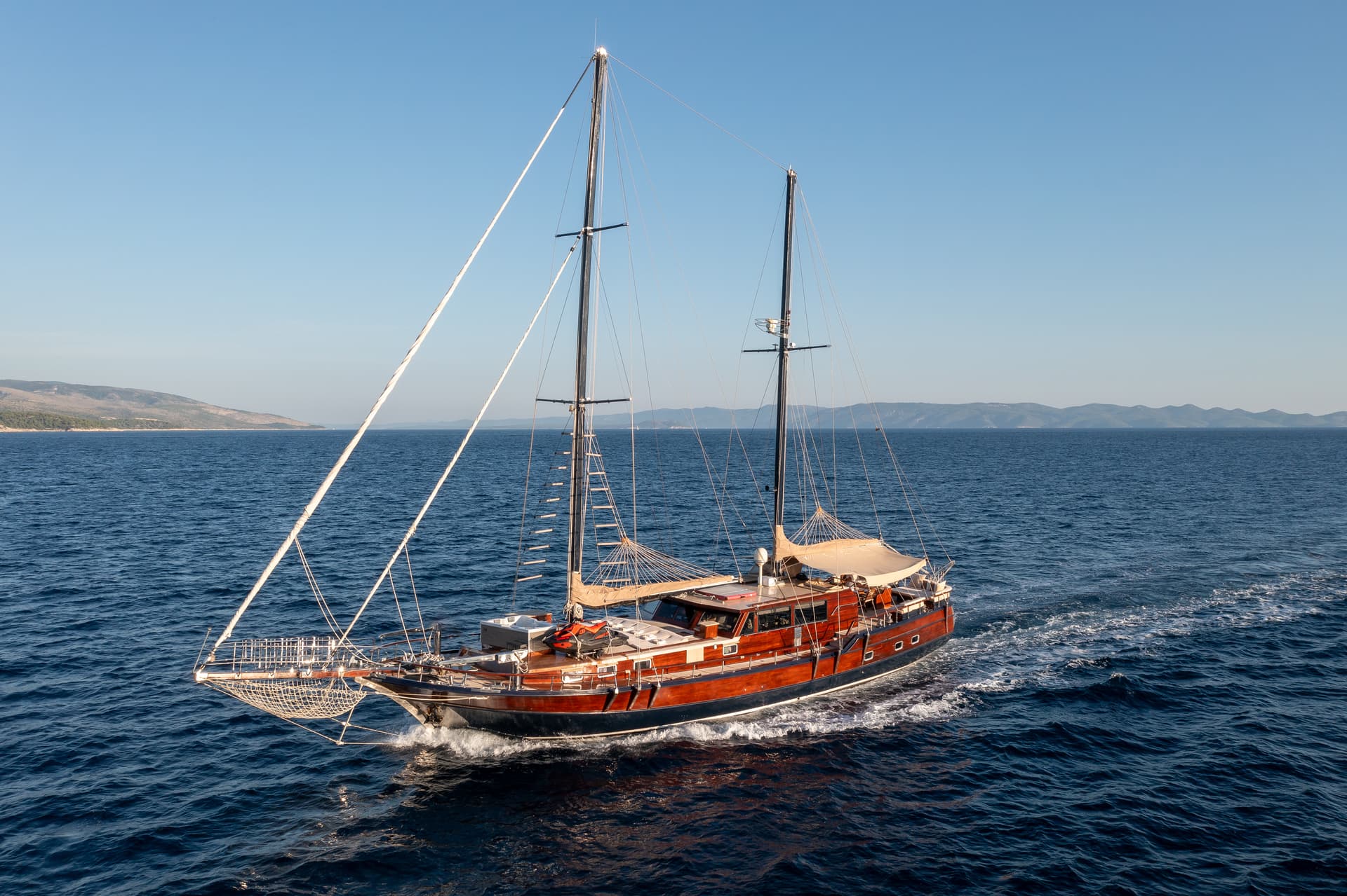 Wooden motor-sailor yacht cruising on deep blue sea near hilly coastline under clear sky