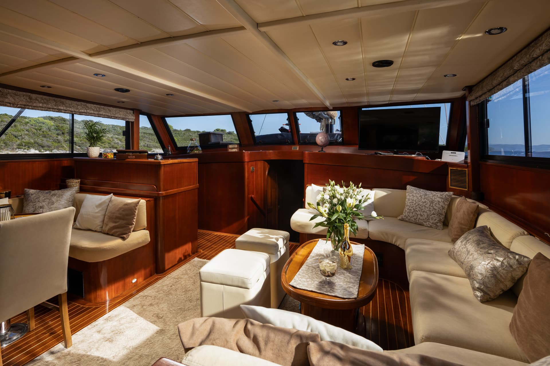 Yacht interior salon with wood trim, beige seating, and view of green coastline.