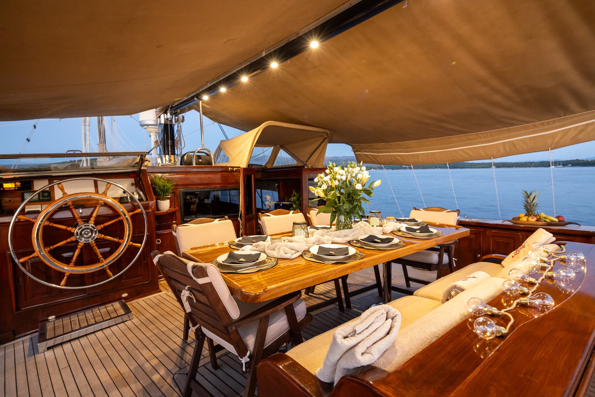 Dining table set on a yacht deck with wooden wheel, overlooking calm blue water at dusk.