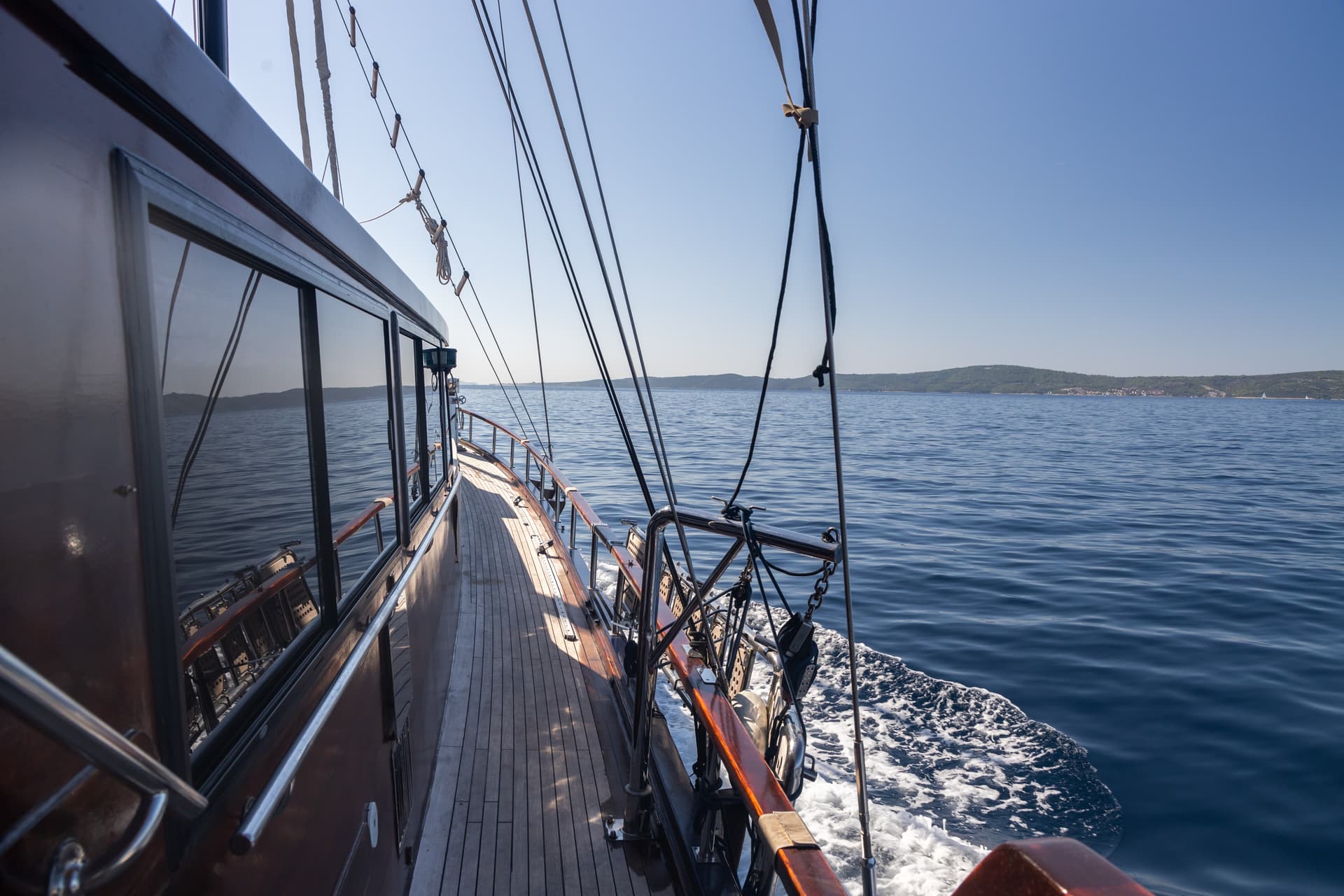 View from wooden boat deck moving on deep blue sea toward green, hilly coastline.
