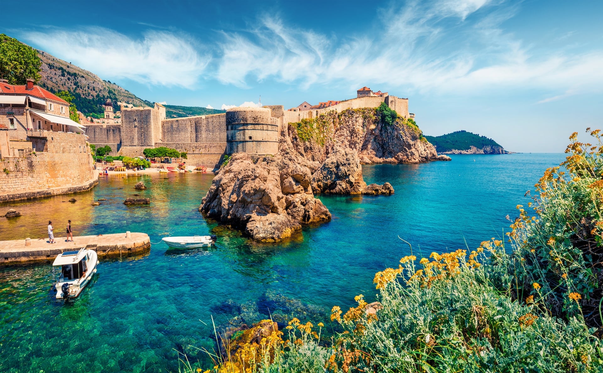 Dubrovnik walls overlooking clear turquoise sea with boats near rocky outcrop and yellow flowers.