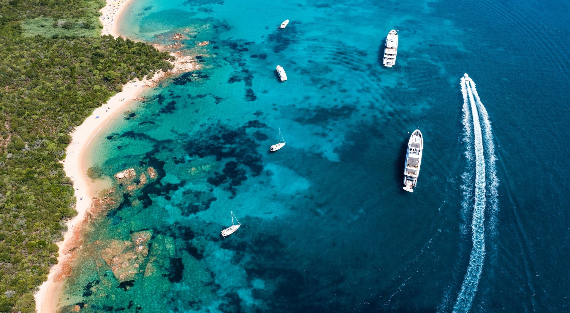 Yachts and sailboats anchored near a lush green coastline with turquoise and deep blue water.