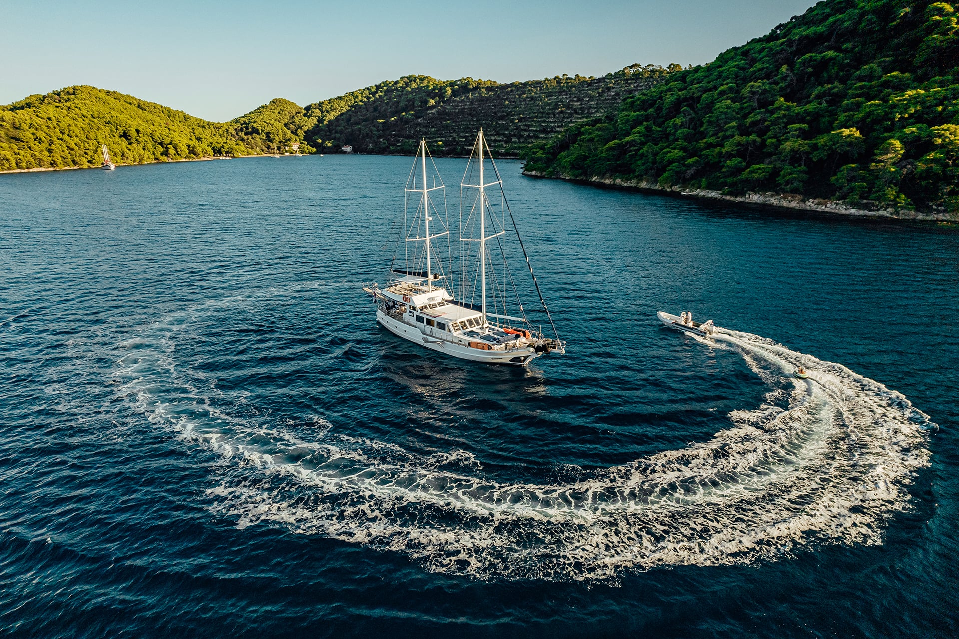 Gulet sailboat anchored near forested coastline while a tender tows an inflatable in dark blue water.