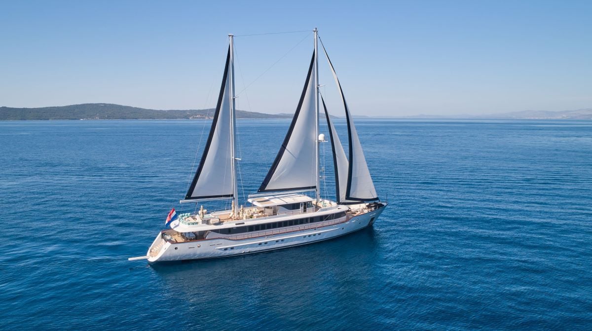 Large white sailing yacht with black-trimmed sails on deep blue sea near hilly coastline.