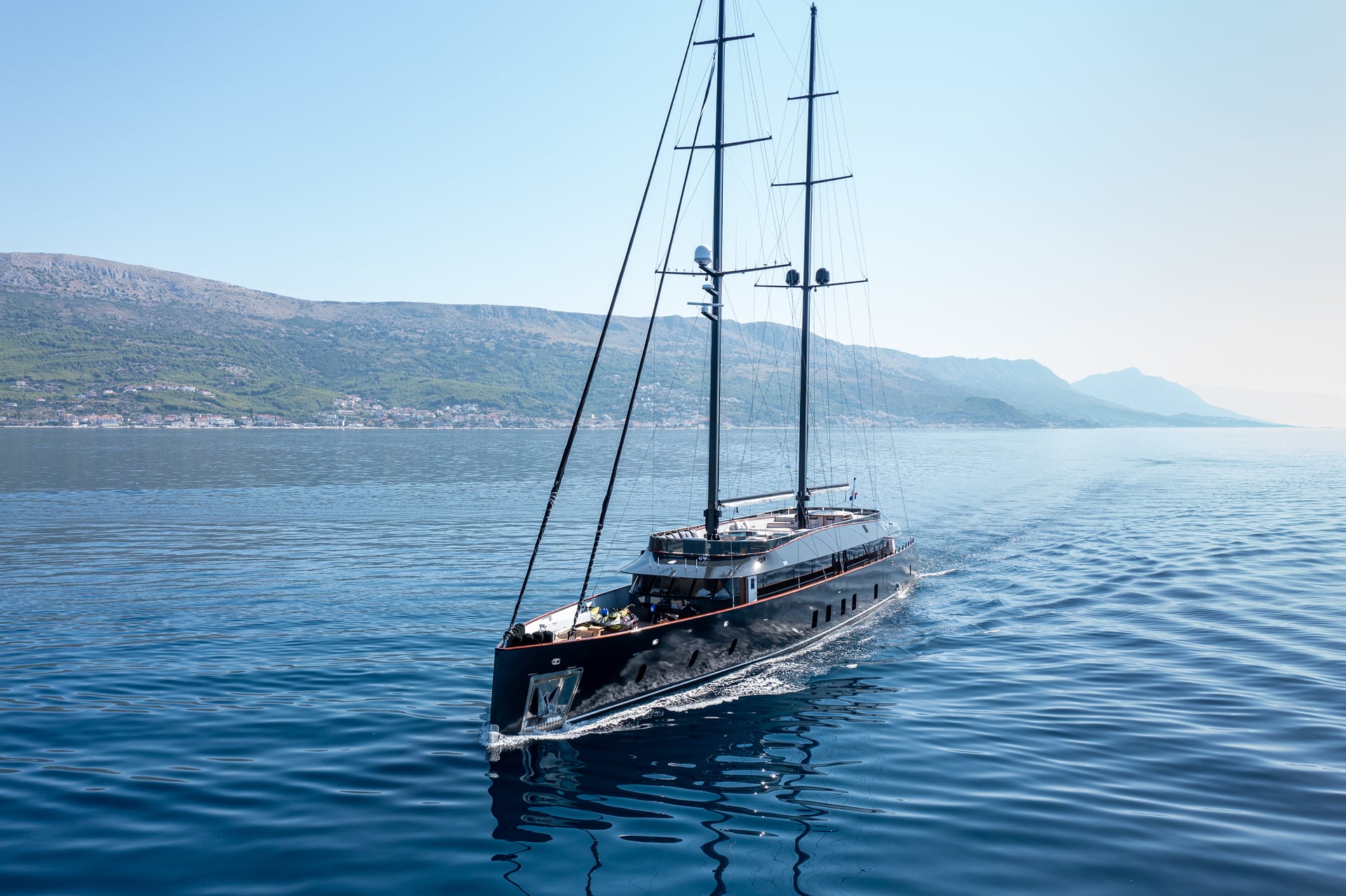 Large black sailing yacht cruising on blue water near a coastal mountain town.