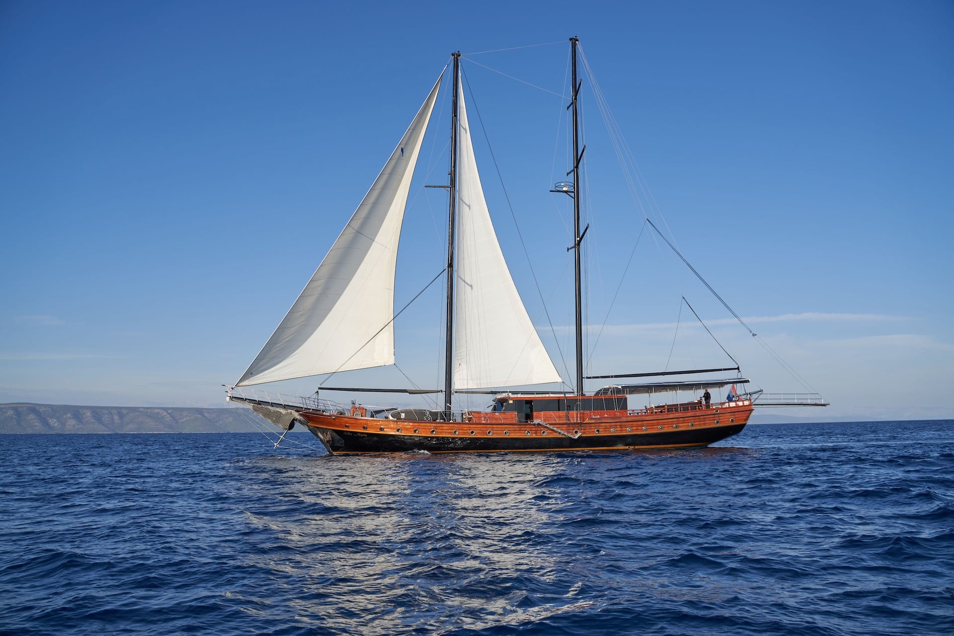Wooden sailboat Stella Maris with white sails on deep blue, choppy sea near distant coastline.