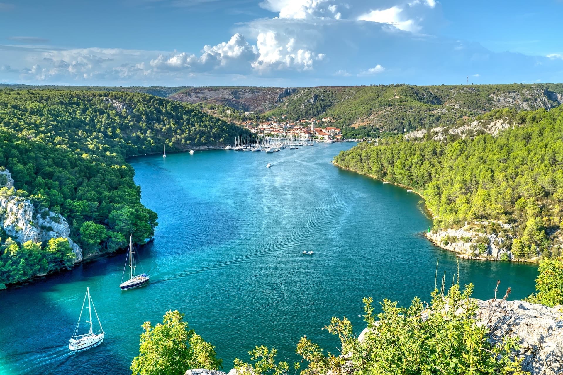 Sailboats on turquoise water inlet with forested hills and Skadrin town in distance.