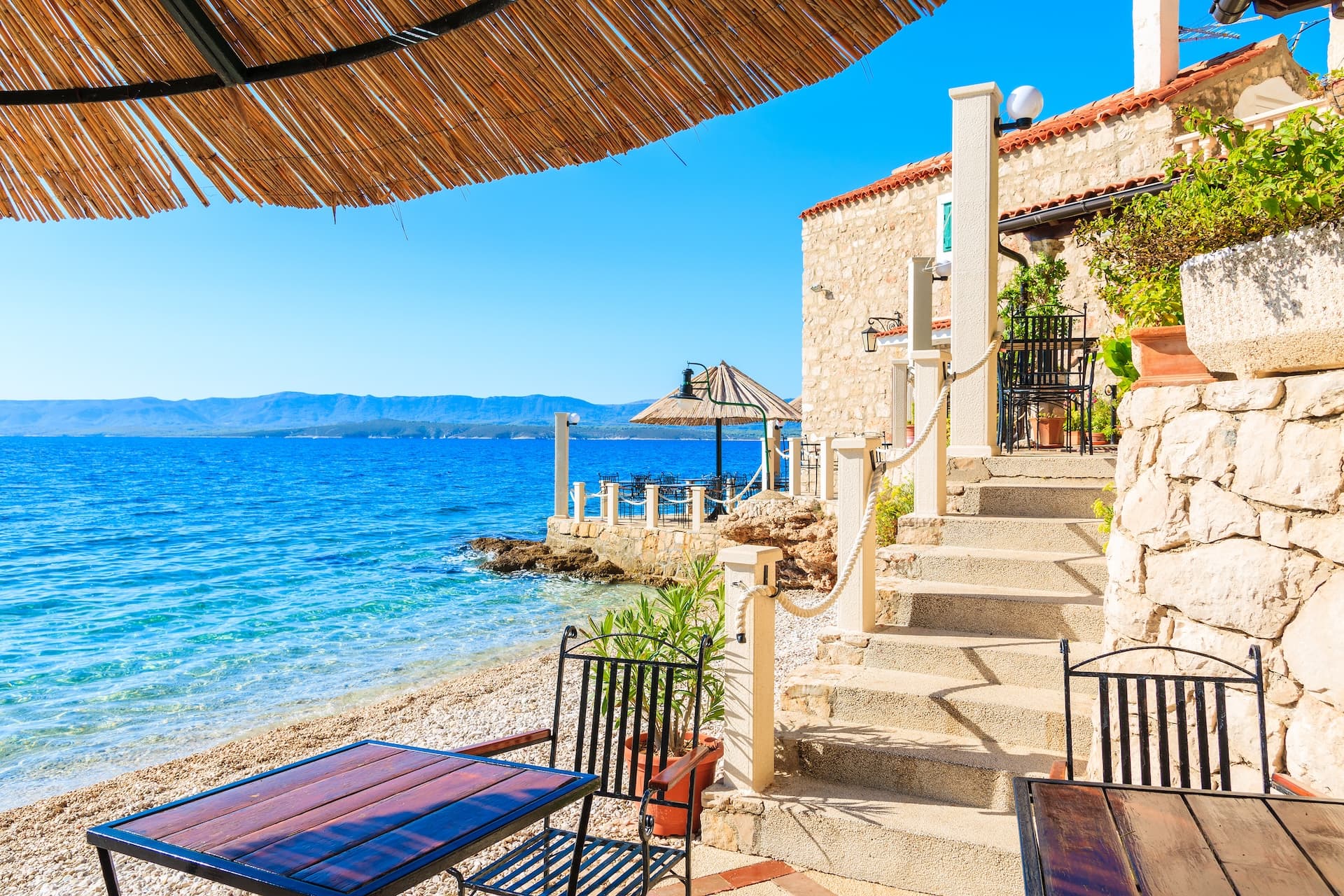 Outdoor cafe seating by pebble beach overlooking blue sea and distant mountains