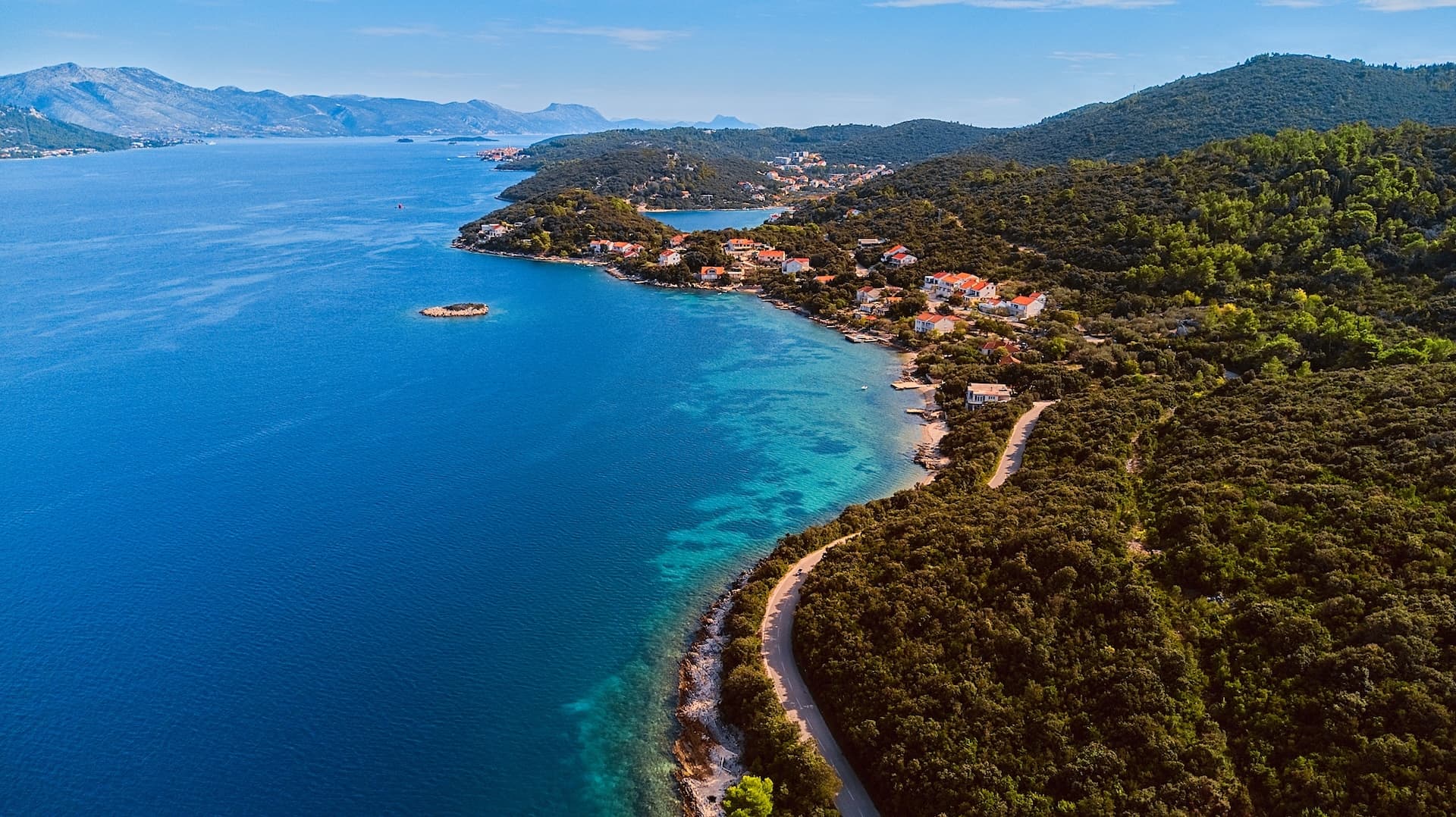 Aerial view of Korcula Island coastline with blue sea, dense green hills, and white houses.