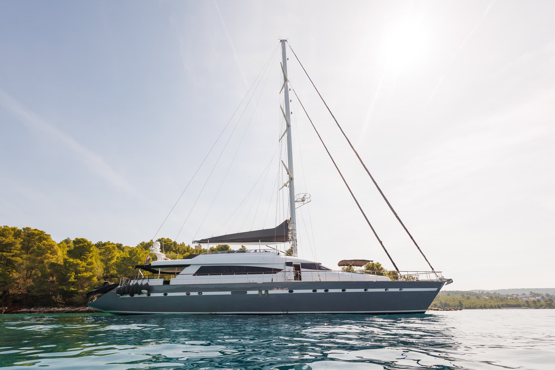 Large sailboat anchored near a lush green coastline under bright sun.