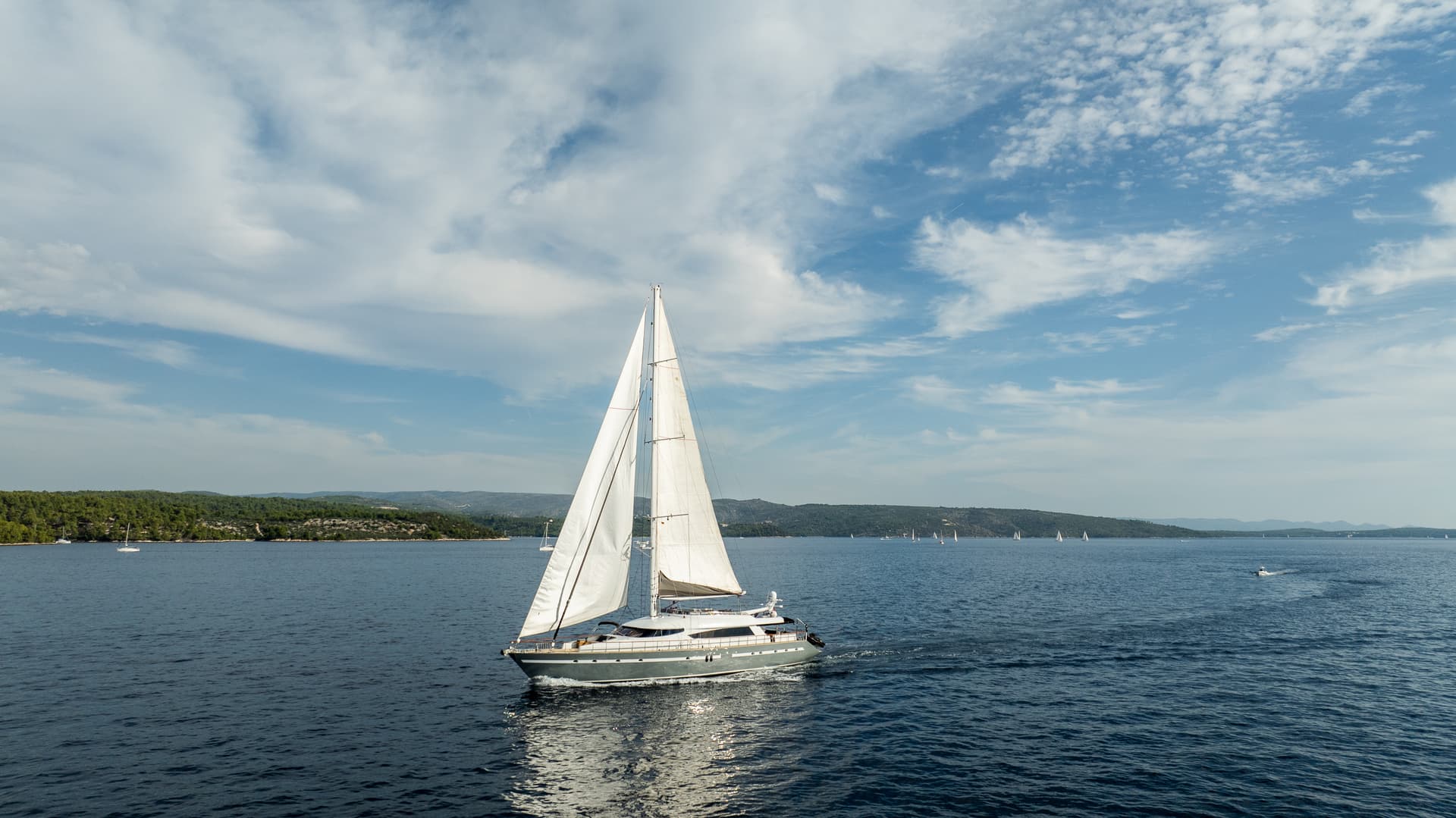 Sailboat cruising on dark blue water near a forested coastline under a partly cloudy sky.