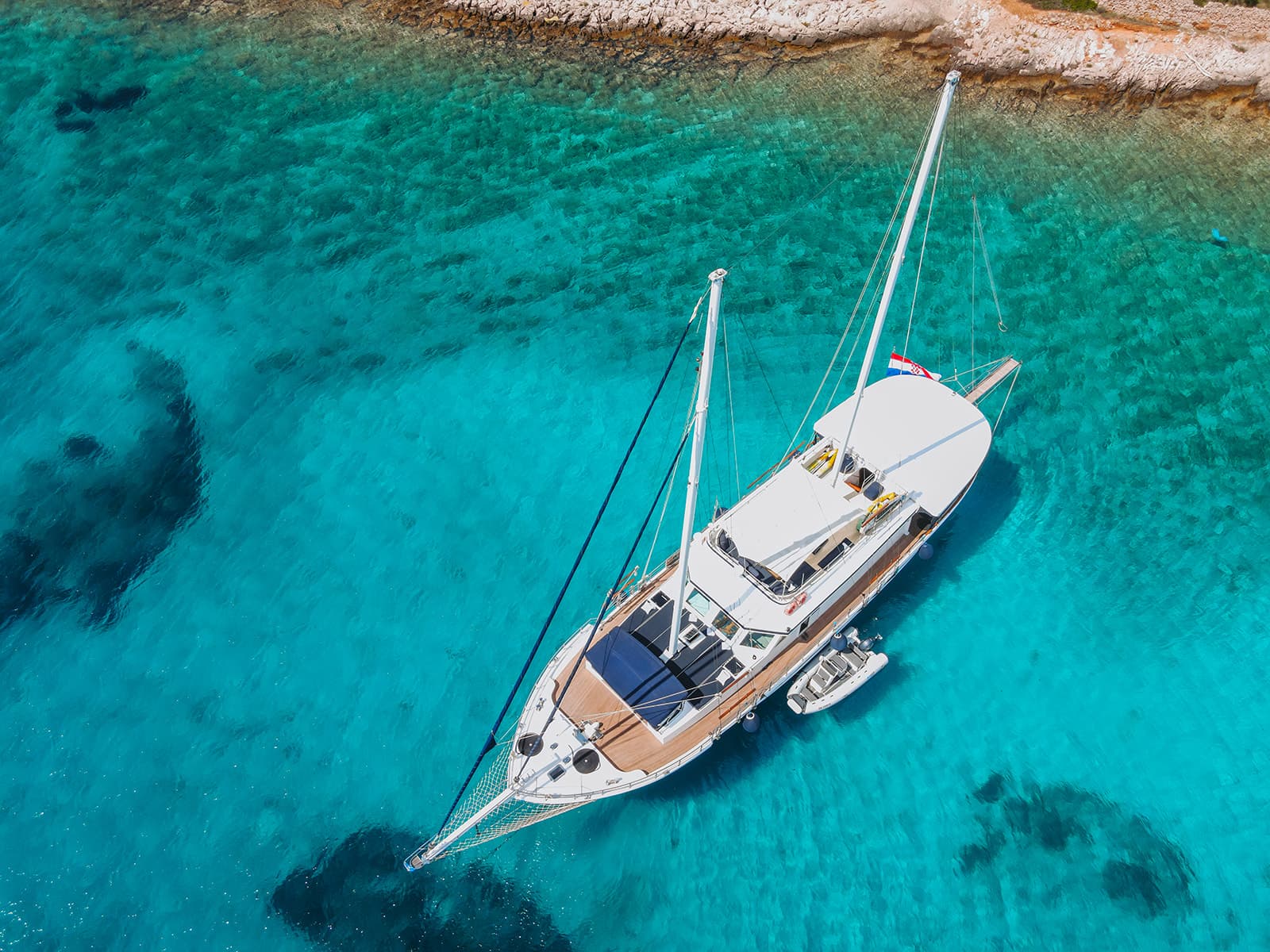 Large white sailboat anchored in clear turquoise coastal water near rocky shore