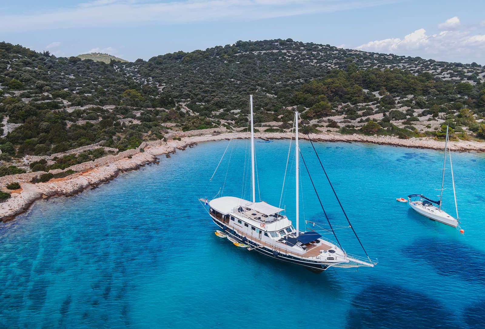 Large sailboat and smaller yacht anchored in turquoise coastal cove near rocky, scrub-covered island.